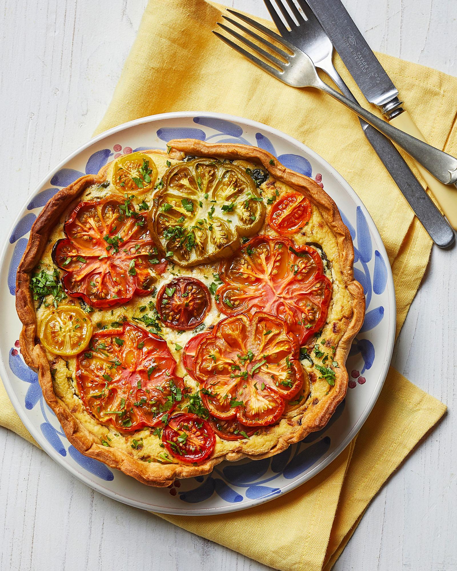 A savory tart with a flaky crust, topped with a mixture of cheese and vegetables, including tomatoes, onions, and herbs, served on a white plate on a yellow napkin.