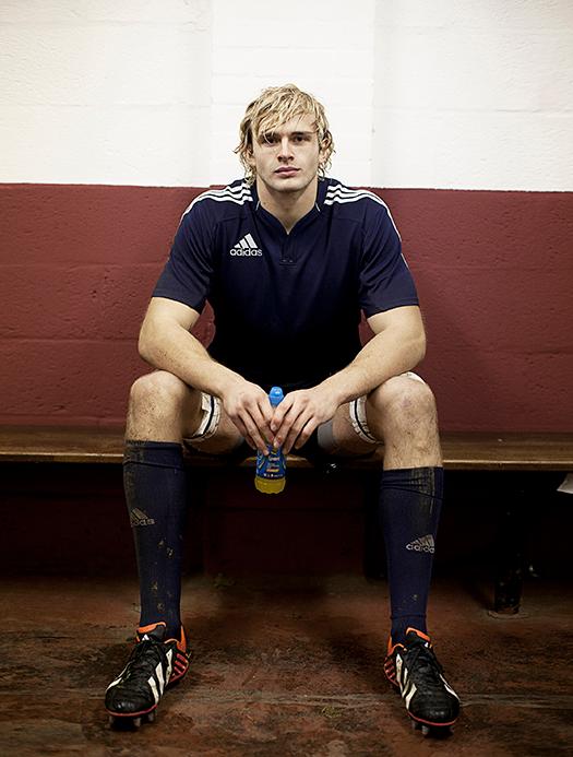 A man in a blue Adidas rugby jersey, sitting on a bench, holding a water bottle.