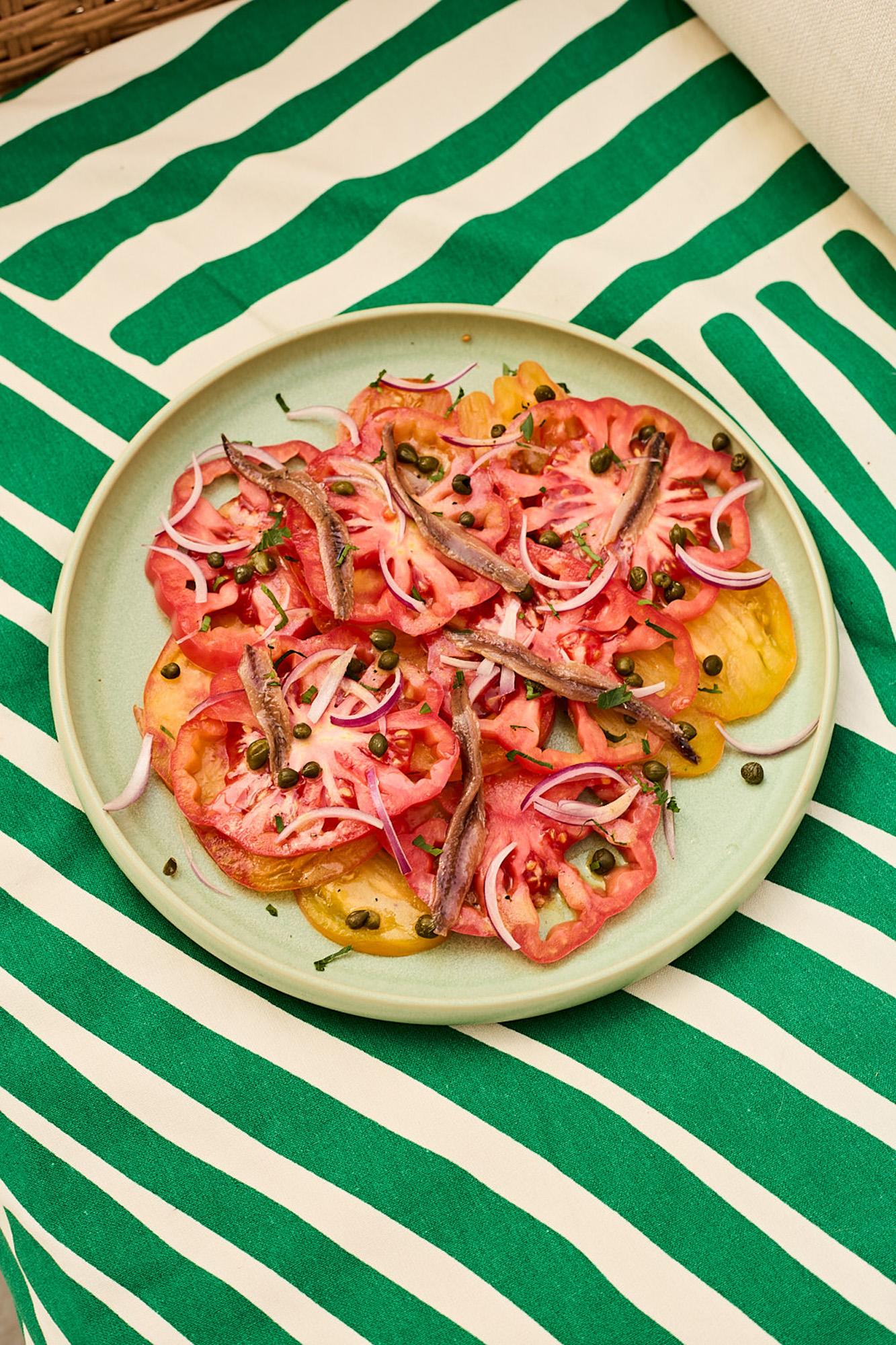 A plate of sliced tomatoes, topped with anchovies, capers, and red onion, served on a tablecloth with a green and white striped pattern.