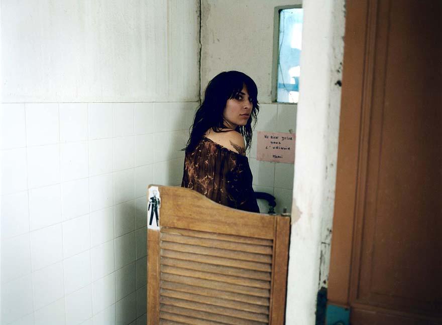 A woman with long, dark hair and a brown, off-the-shoulder top stands in a dimly lit, white-tiled room, looking over her shoulder.