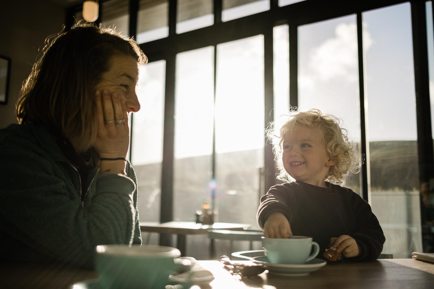 A woman and a young child sitting at a table in a cafe, both smiling and laughing together.