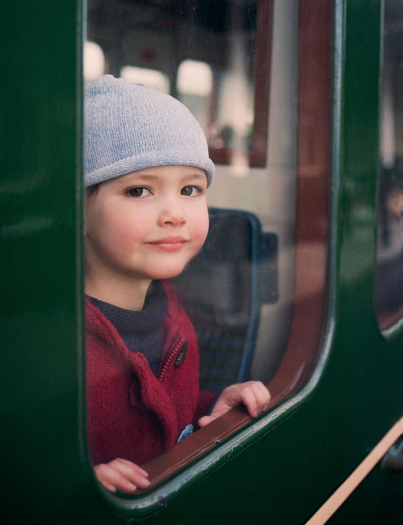 A young boy wearing a red fleece jacket and a grey beanie, looking out of a train window.