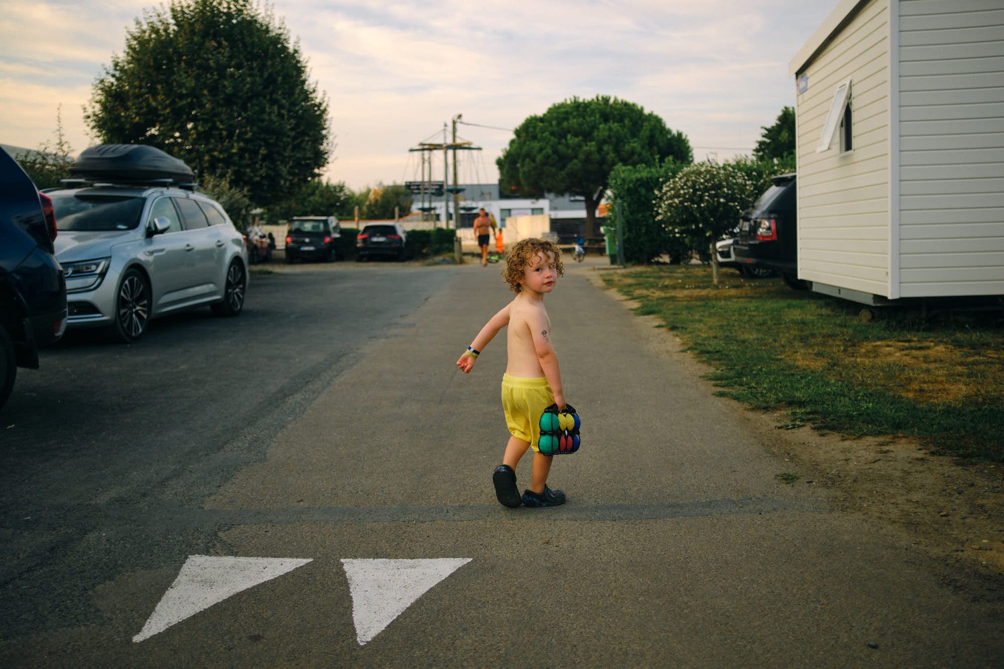 A young boy, shirtless and with curly hair, walks across a road, holding a colorful toy. He appears to be in a campsite or car park, surrounded by cars and trees.