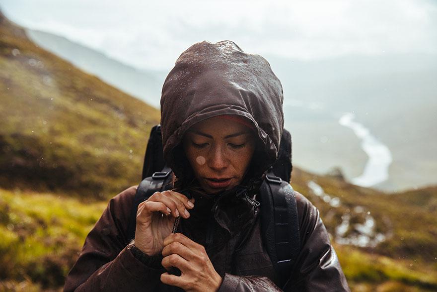A woman in a brown waterproof jacket and backpack, hiking in the rain, with a mountainous landscape in the background.