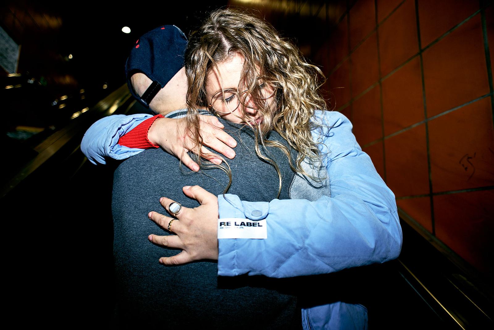 A woman in a blue jacket and a man in a grey sweater embracing in a dimly lit, urban setting.