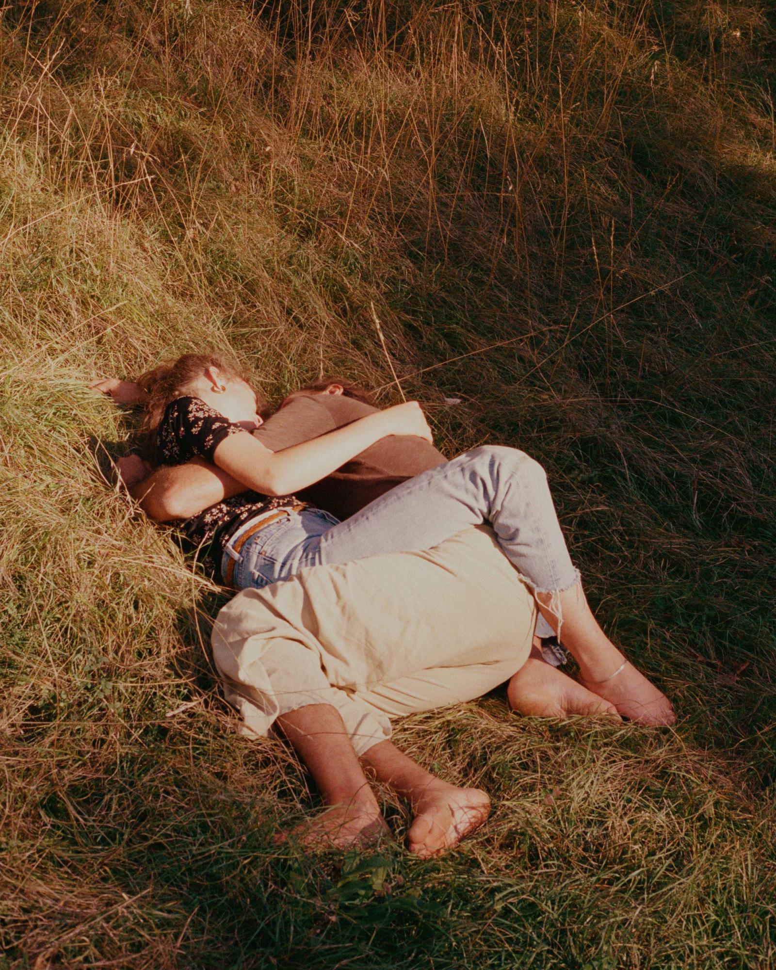 A couple is embracing and kissing in a field of tall, dry grass, with the woman wearing a black floral blouse and the man wearing a brown shirt and jeans.
