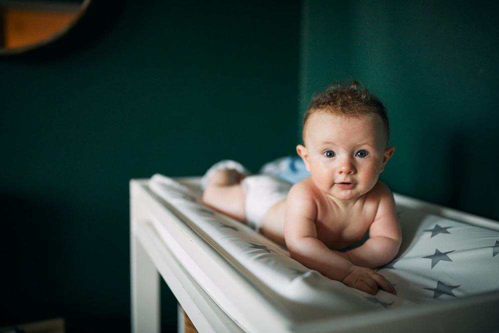 A baby lies on a changing table, looking directly at the camera with a curious expression. The baby is naked, with a diaper on, and is lying on a white changing table with a.