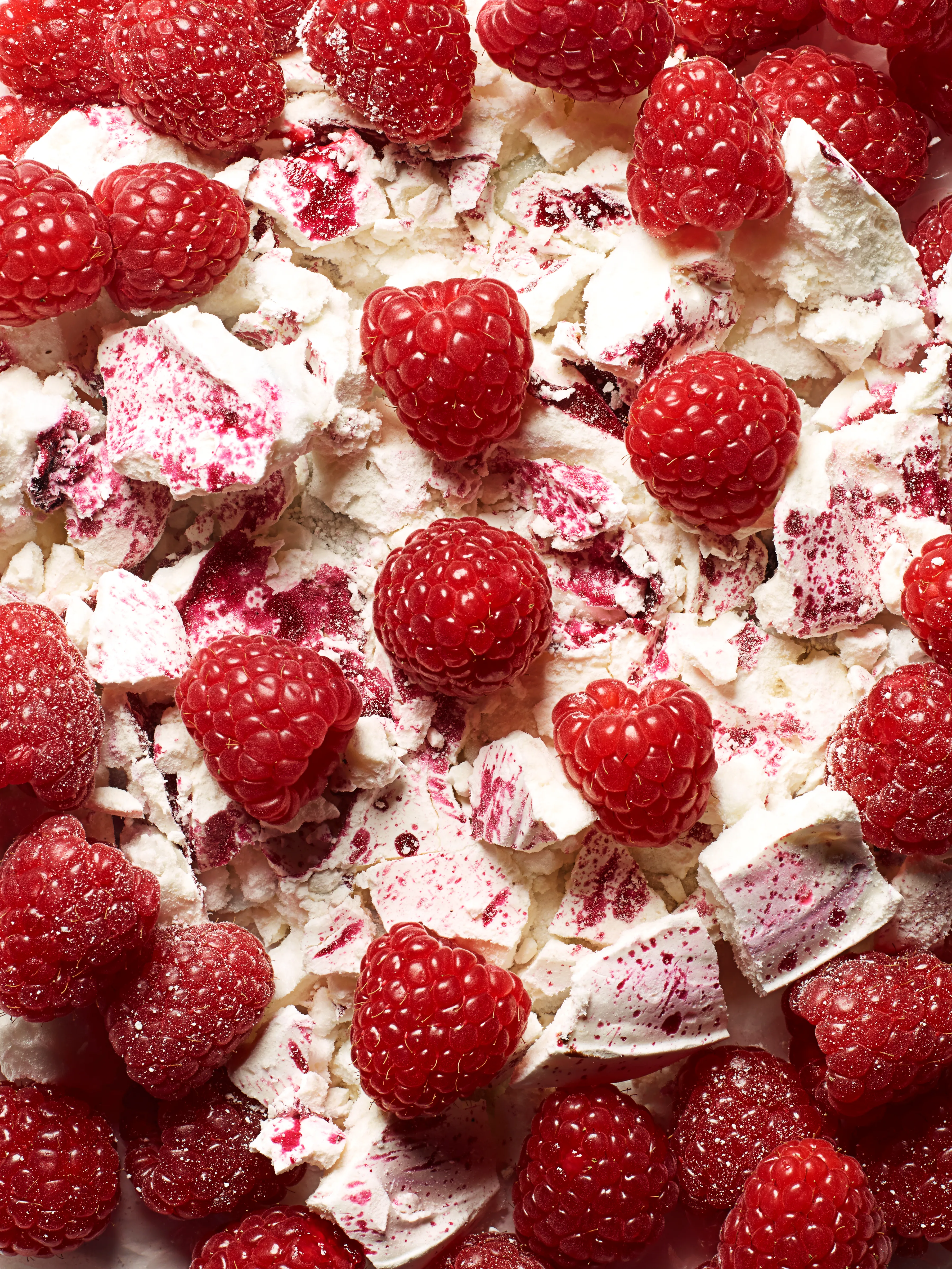 A close-up of a large, white, meringue-like dessert with a raspberry topping, with many fresh raspberries scattered on top.