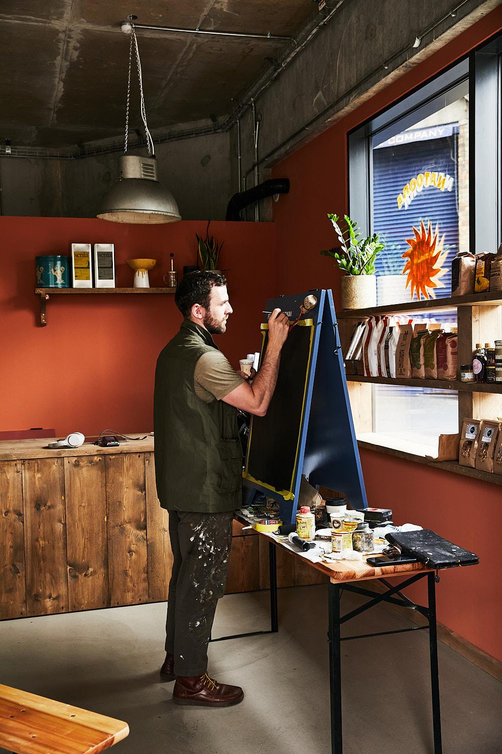 A man in a green vest and paint-splattered pants stands in a studio, holding a large, blue, A-frame sign with a blackboard surface, surrounded by art supplies and paint.