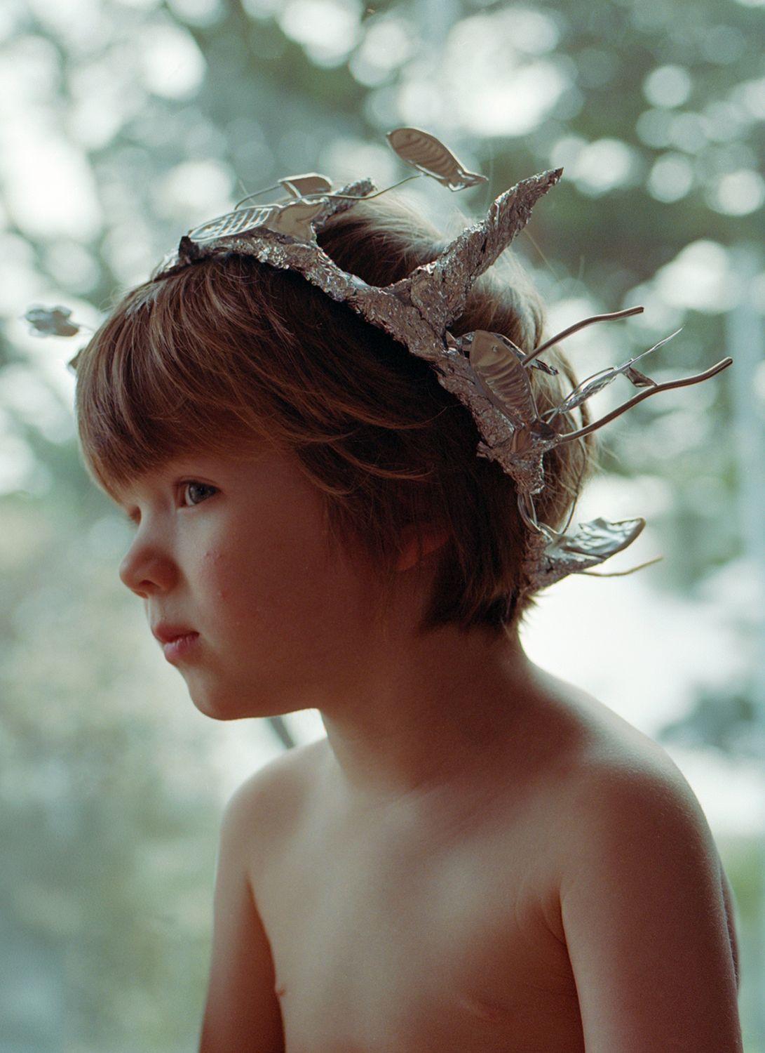 A young boy with a crown of leaves on his head, looking to the left with a serious expression. He has short, brown hair and is shirtless, with his arms at his sides. The.