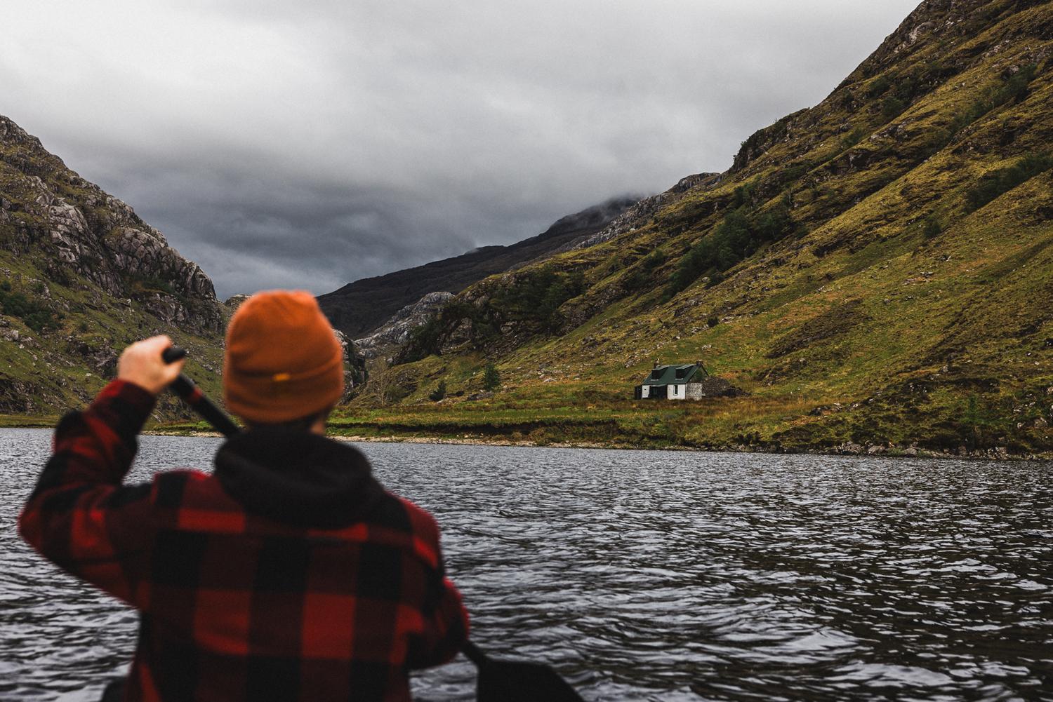 A person in a red and black plaid jacket and an orange beanie, paddling a canoe in a serene mountain lake, with a small cabin in the distance.