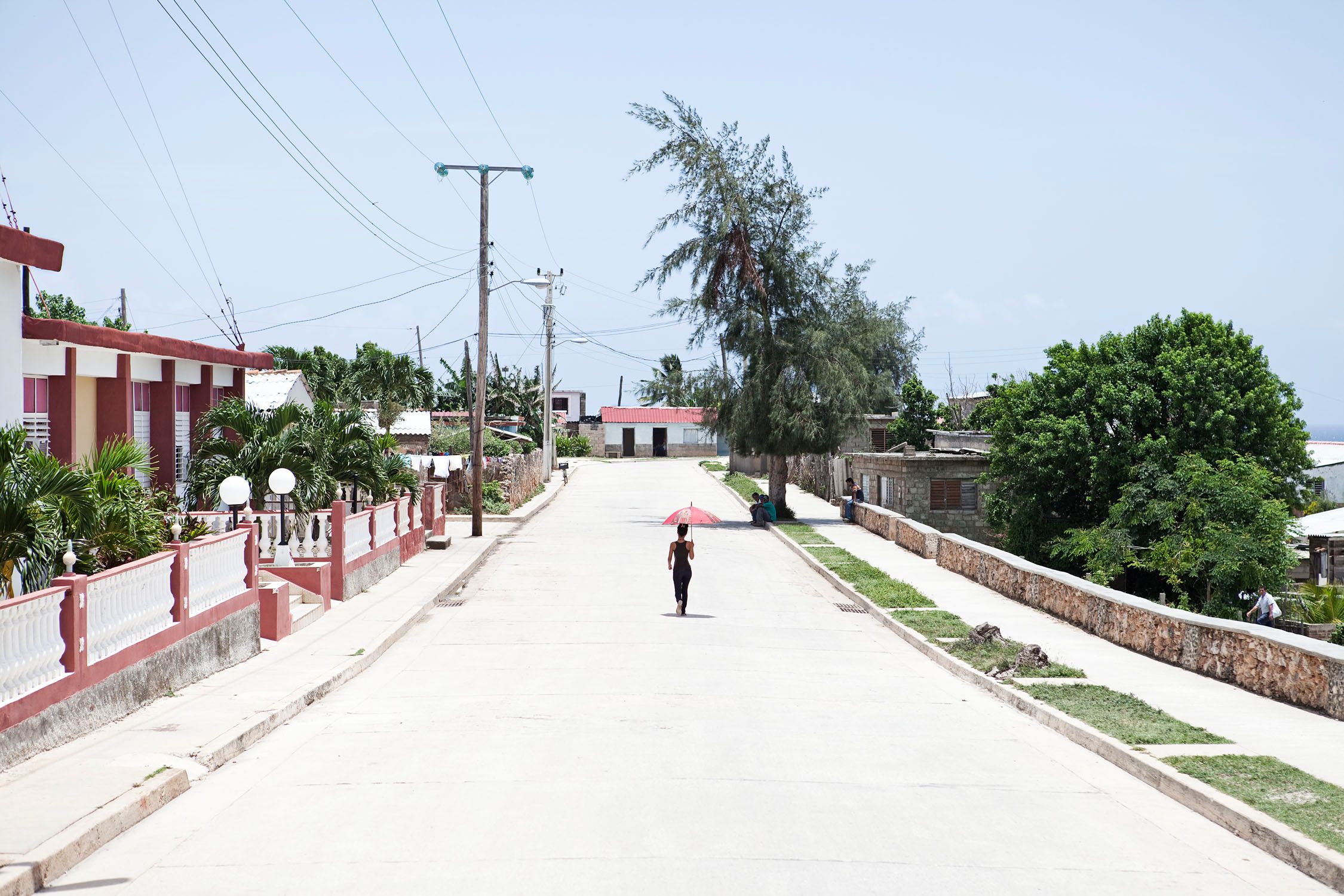 A person walking down a deserted street in a tropical town, holding a red umbrella.