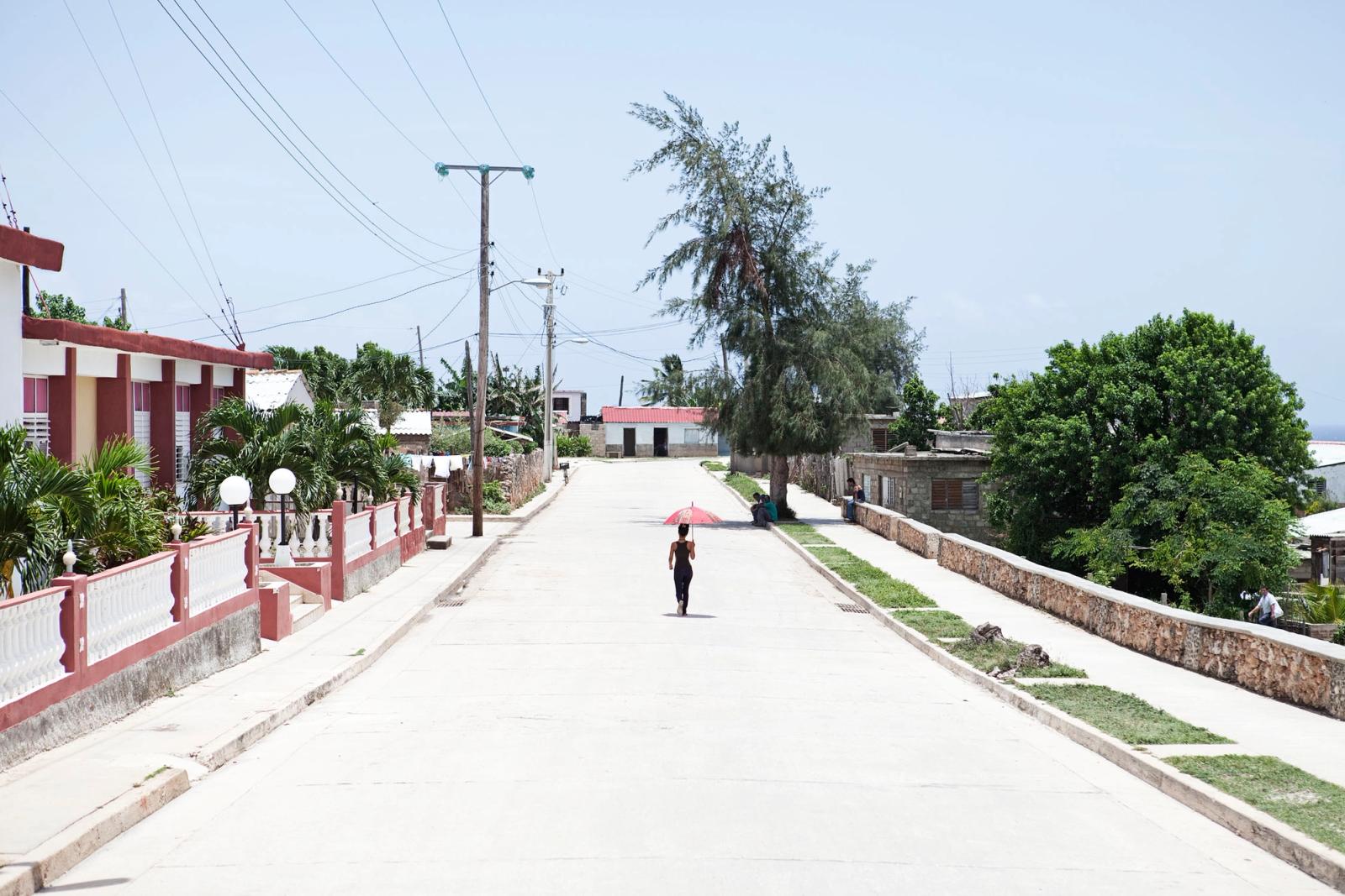A person walking down a deserted street in a tropical town, holding a red umbrella.
