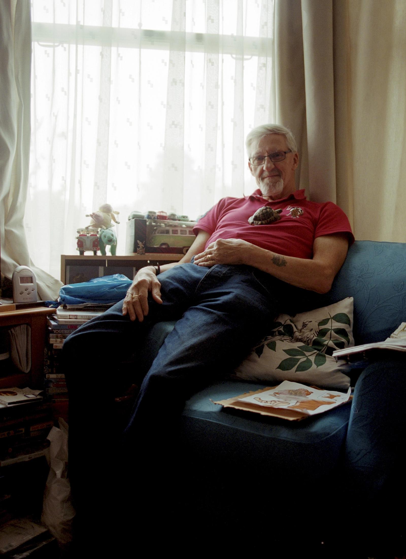 An older man with white hair and a red polo shirt is sitting on a blue couch, surrounded by books and a small, dark-colored animal on his chest.