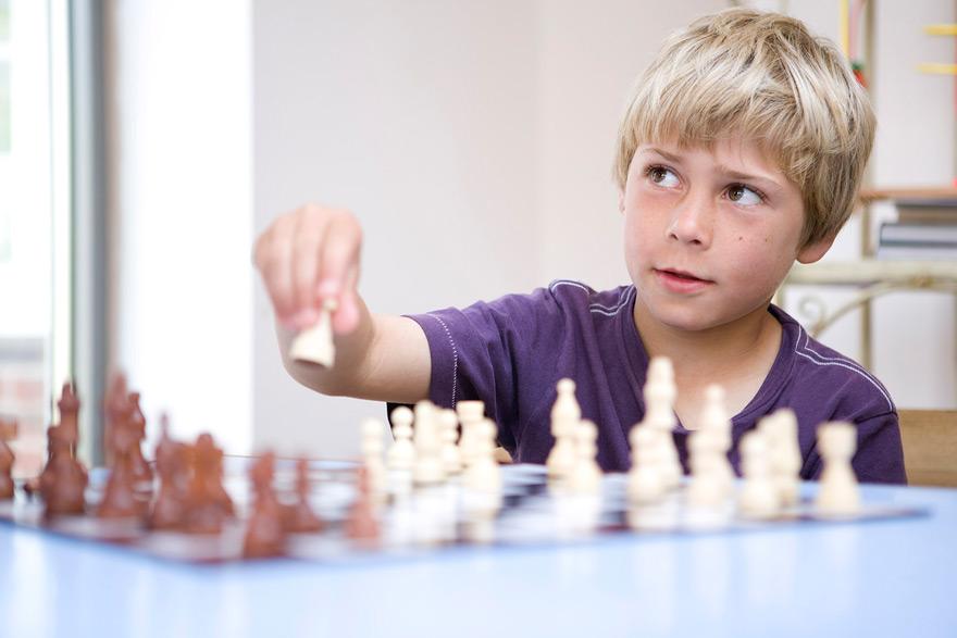 A young boy with blonde hair and a purple t-shirt is playing a game of chess, focusing on the board in front of him.