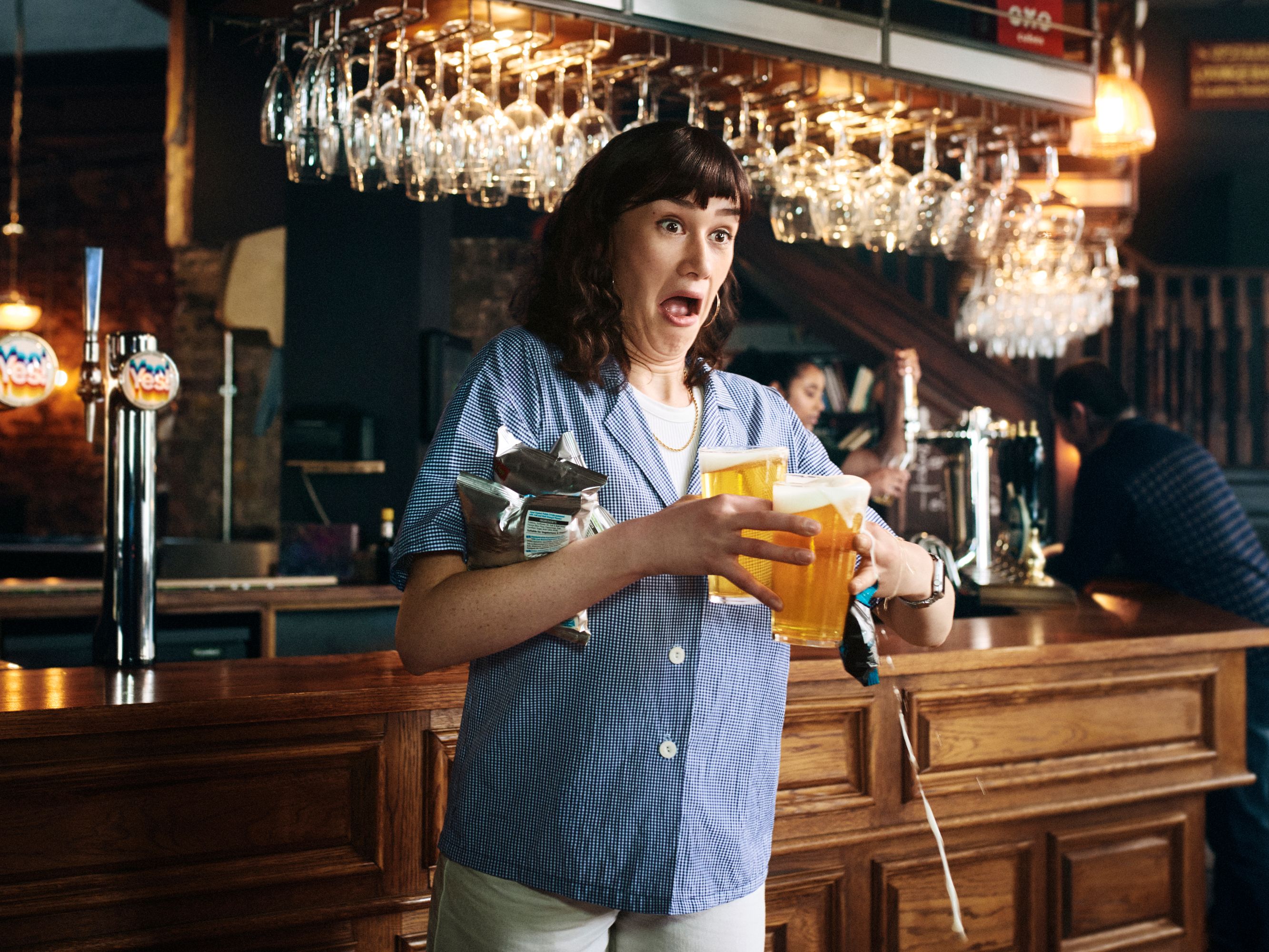 A woman in a blue and white gingham shirt, holding two glasses of beer at a bar, with a surprised expression.