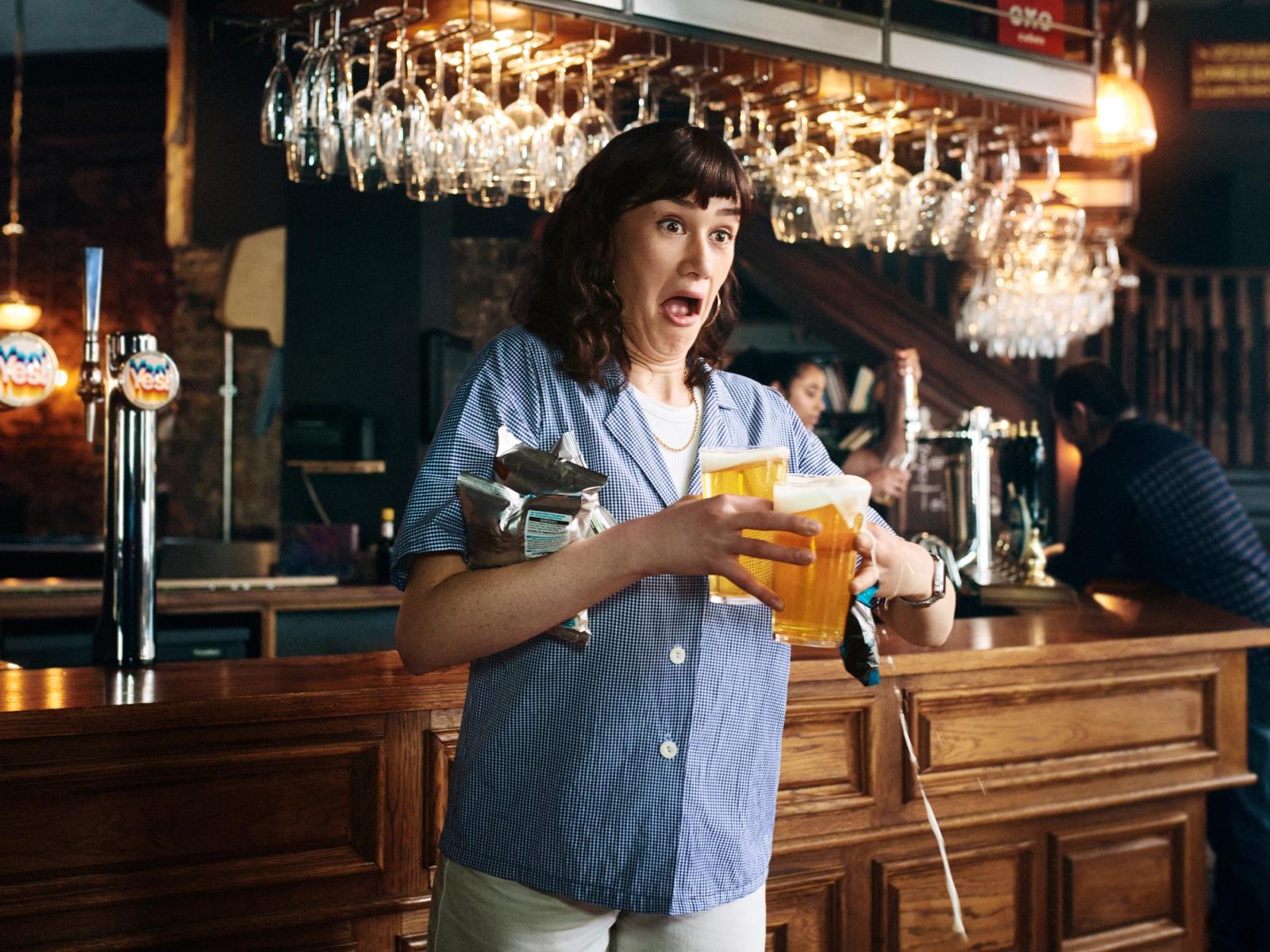 A woman in a blue and white gingham shirt, holding two glasses of beer at a bar, with a surprised expression.