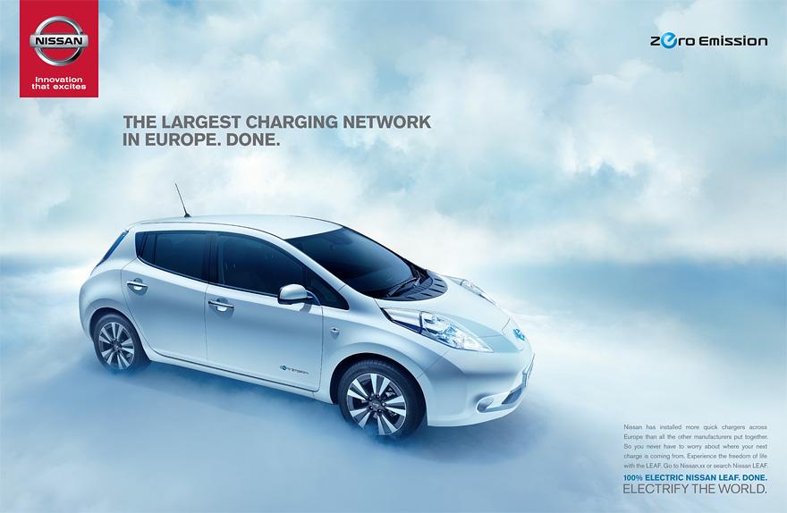 A silver Nissan Leaf electric car is parked in a city street, with a cloudy sky in the background.