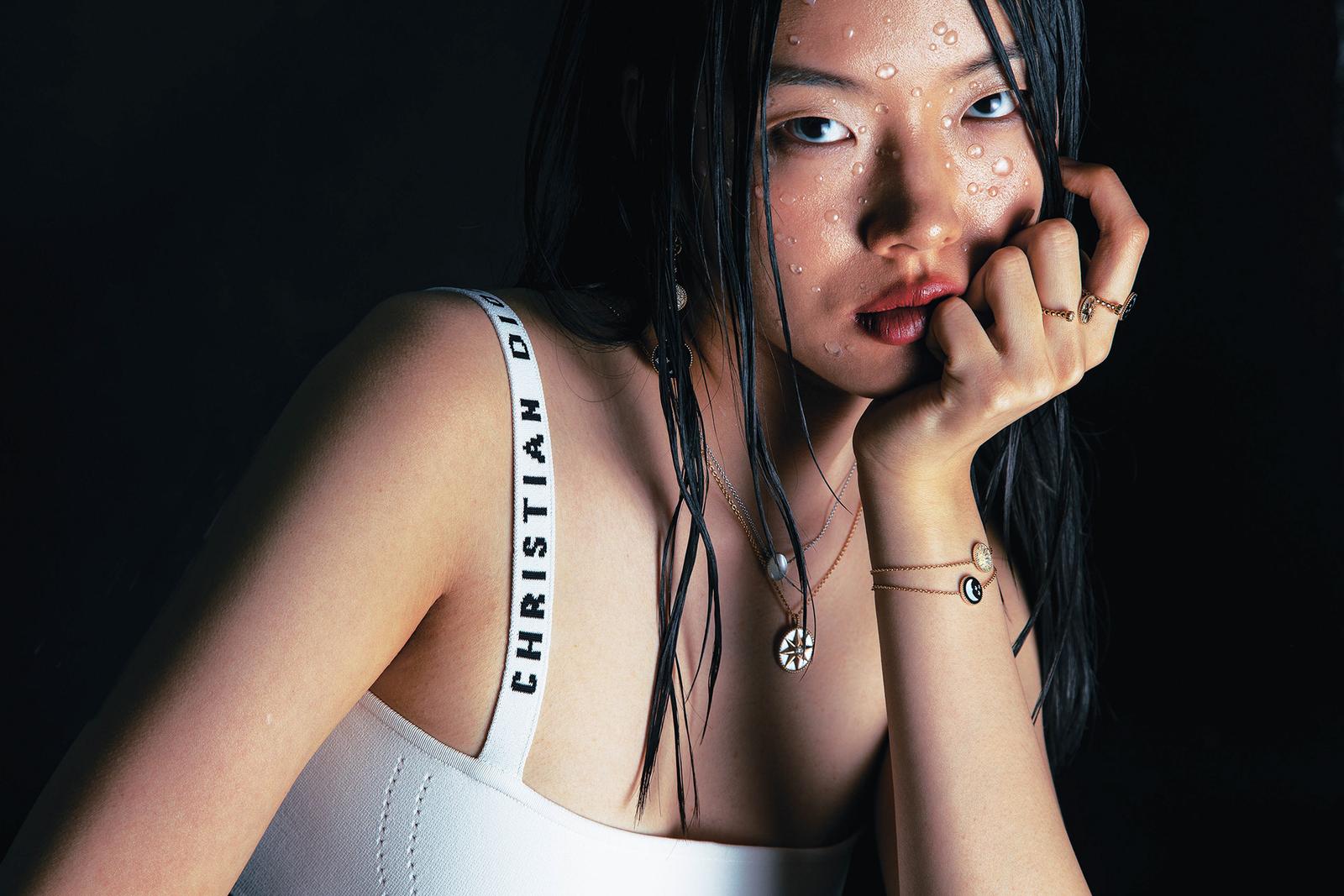 Darina Umka posing in a white tank top with a subtle Christian Dior logo on the strap, with her hand resting on her face and her hair wet and styled in a sleek, straight look.