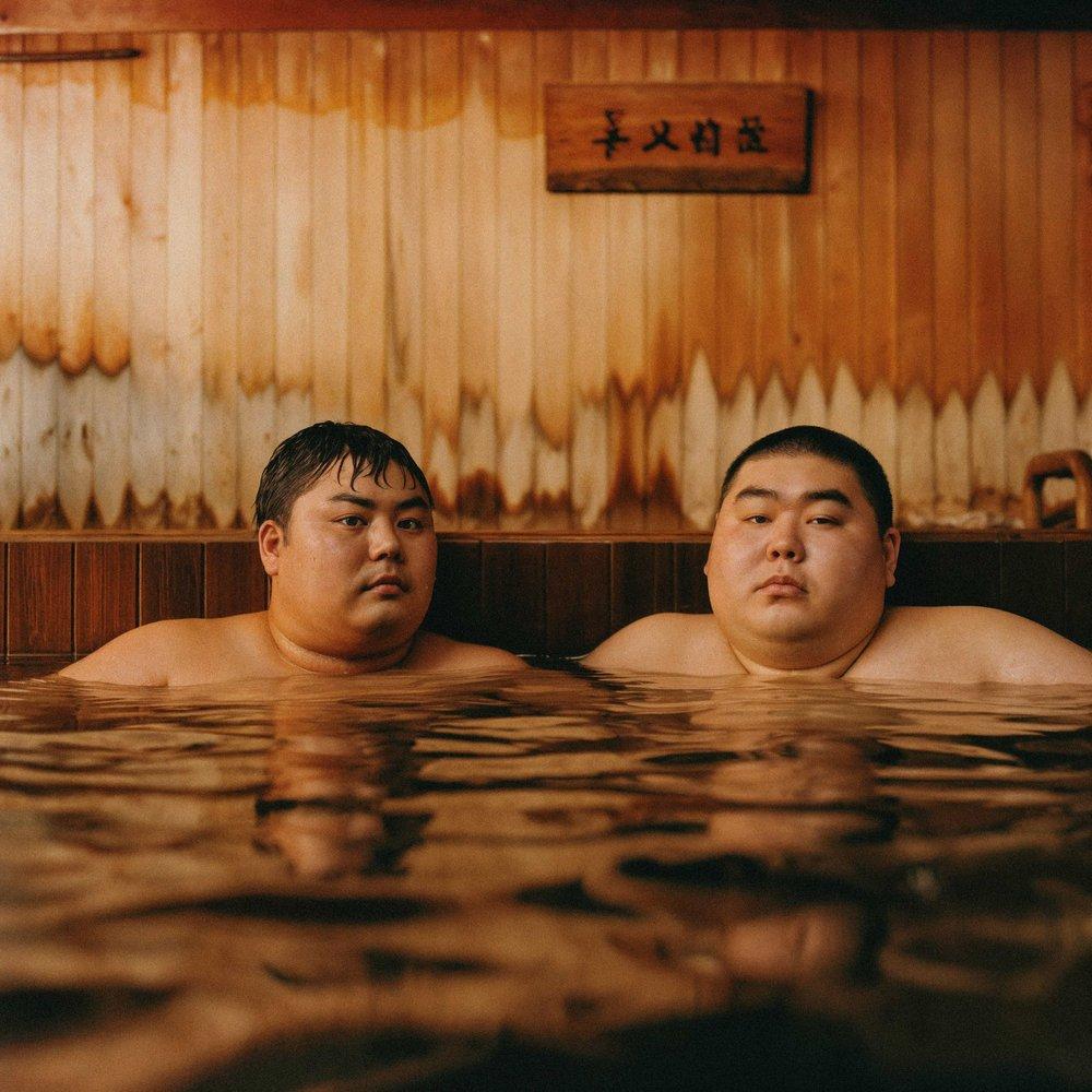Two Asian men in a hot spring or onsen, both with short hair and facial hair, wearing only their underwear or swimwear, in a wooden or traditional Japanese-style setting.
