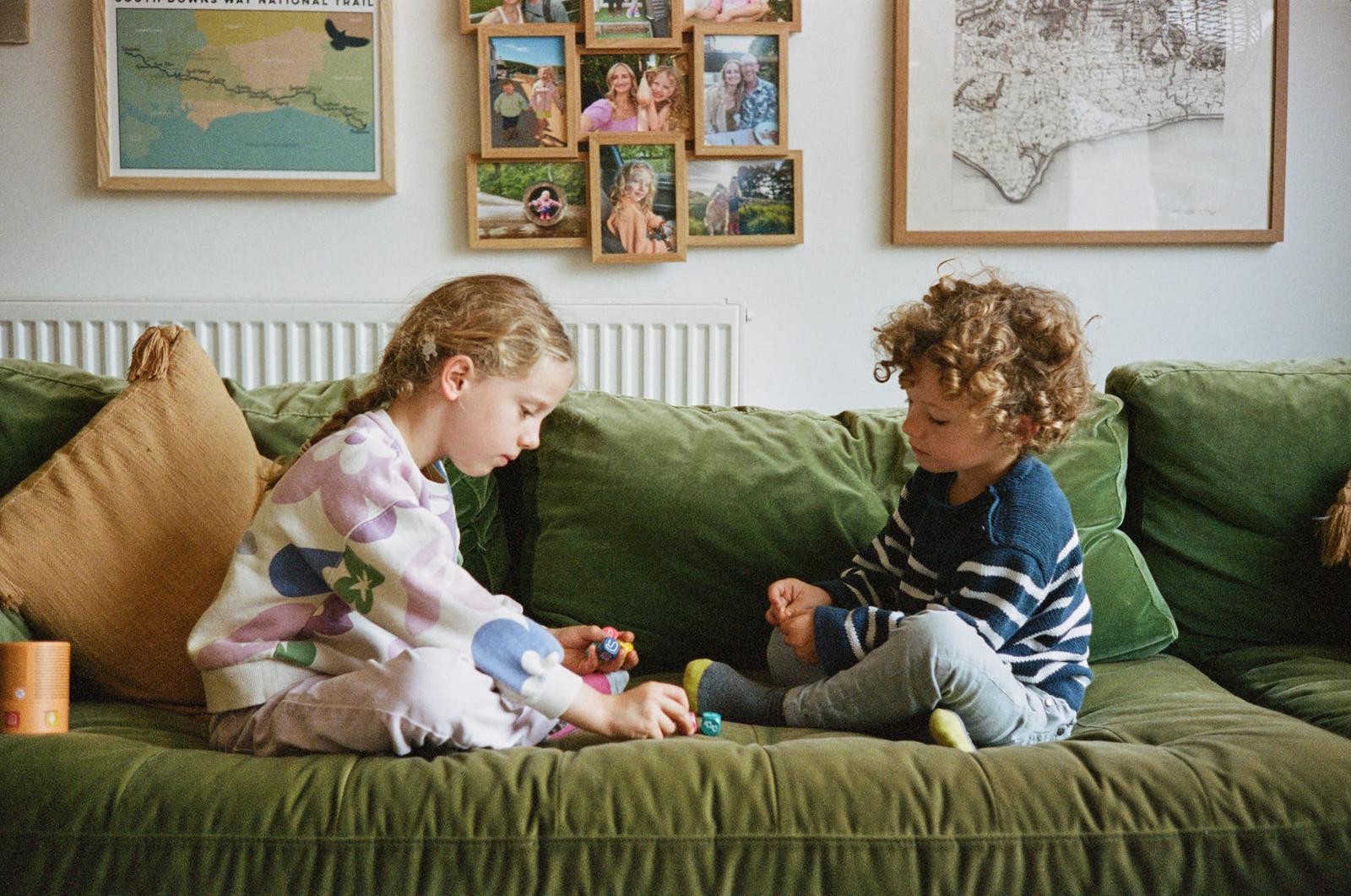 Two children sitting on a green couch, playing with small toys. The girl is wearing a colorful sweater and the boy has a blue and white striped shirt. They both have curly hair.