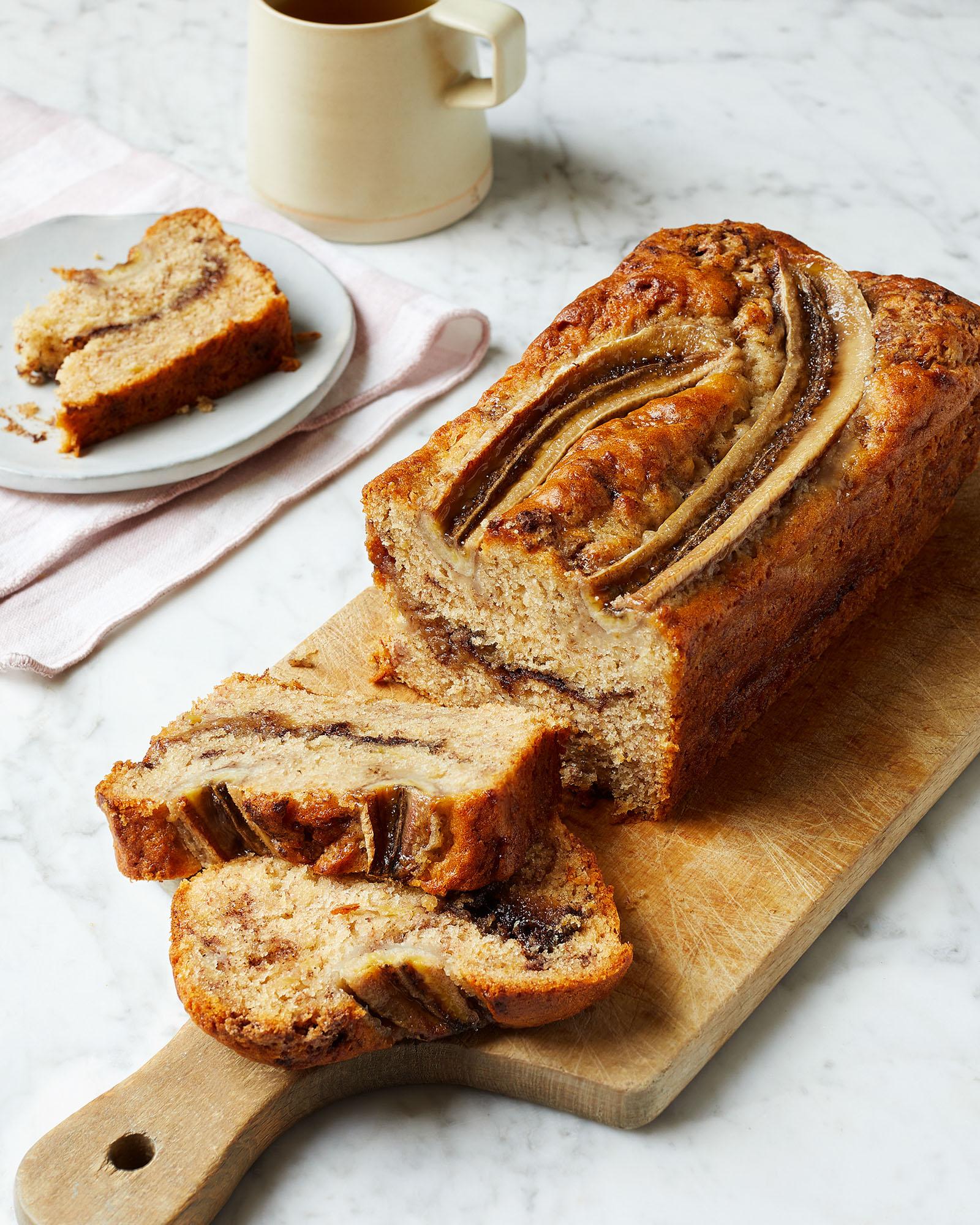A slice of banana bread on a wooden cutting board with coffee nearby.
