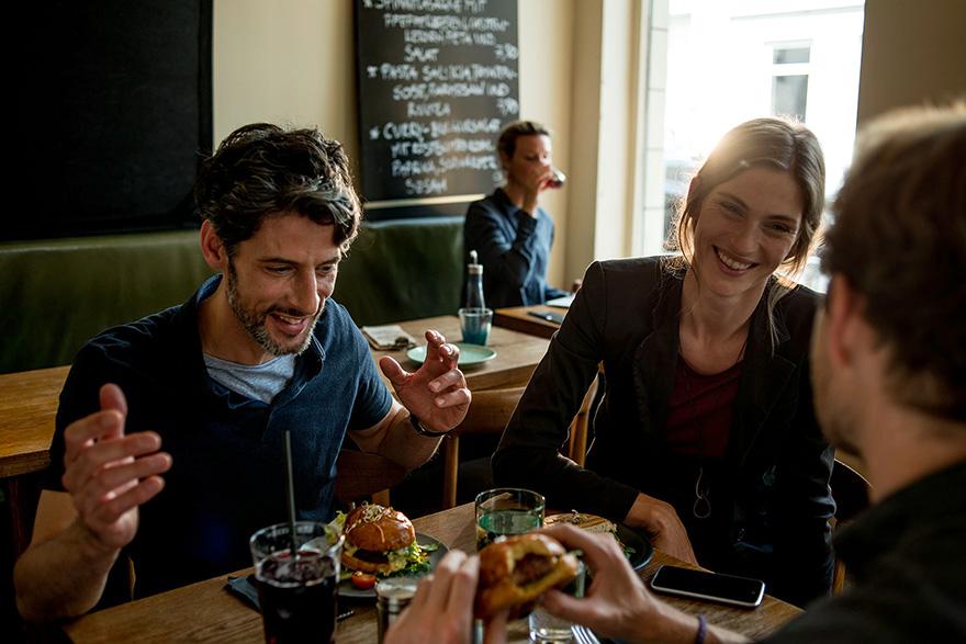 A group of people laughing and eating at a restaurant, with multiple burgers and drinks on the table.