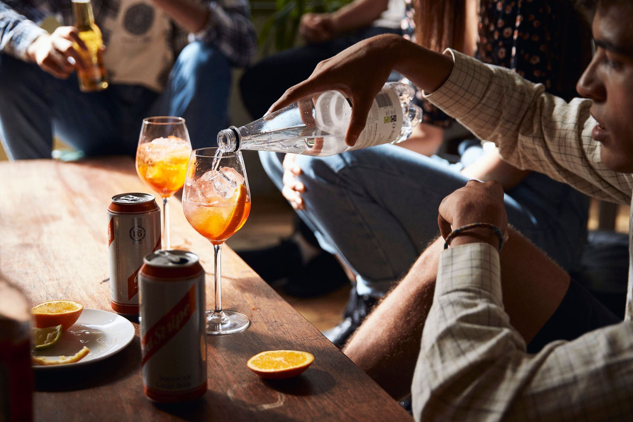 A man in a white shirt pours a drink from a bottle into a glass, surrounded by two other glasses, a plate with an orange slice, and a can of beer. The atmosphere appears to be a.