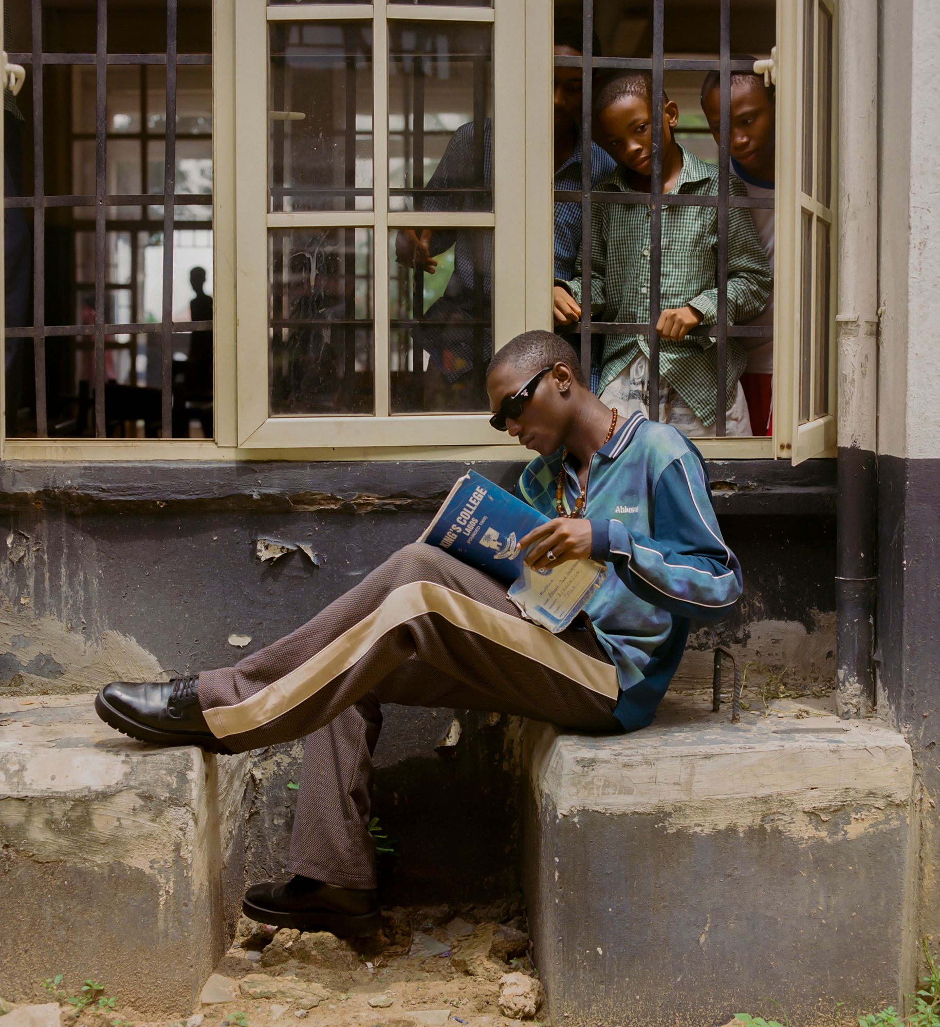 A young man in a blue and white striped shirt, brown pants, and black shoes is sitting on a concrete step, reading a book.
