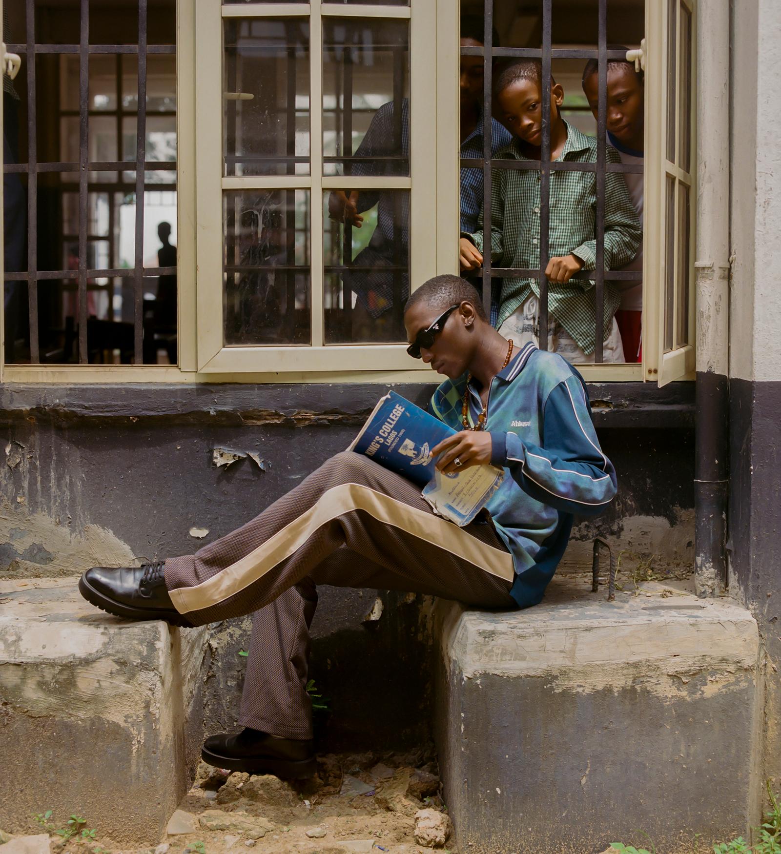 A young man in a blue and white striped shirt, brown pants, and black shoes is sitting on a concrete step, reading a book.