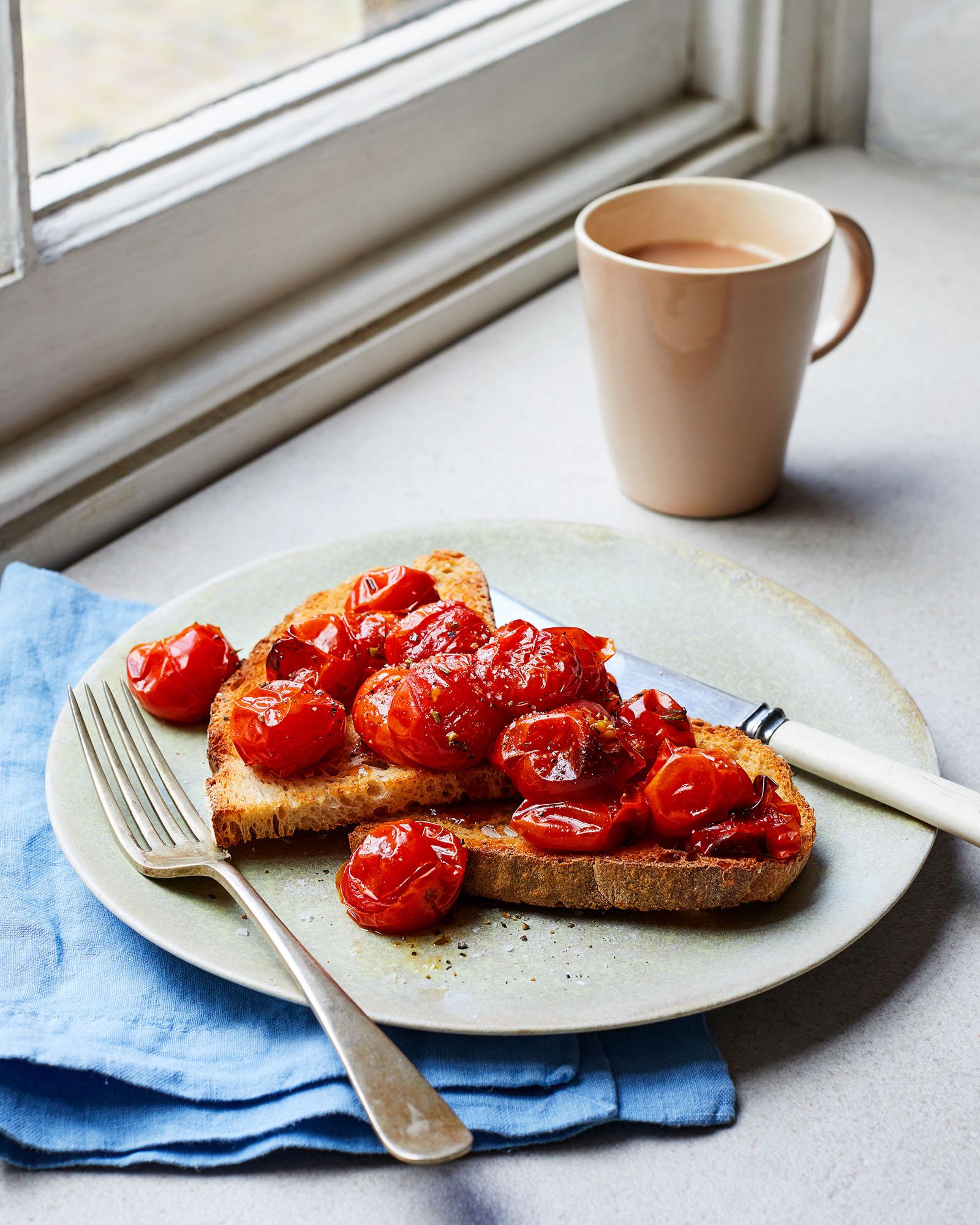 A plate of toasted bread topped with cherry tomatoes, served with a fork and a cup of coffee on a table by a window.