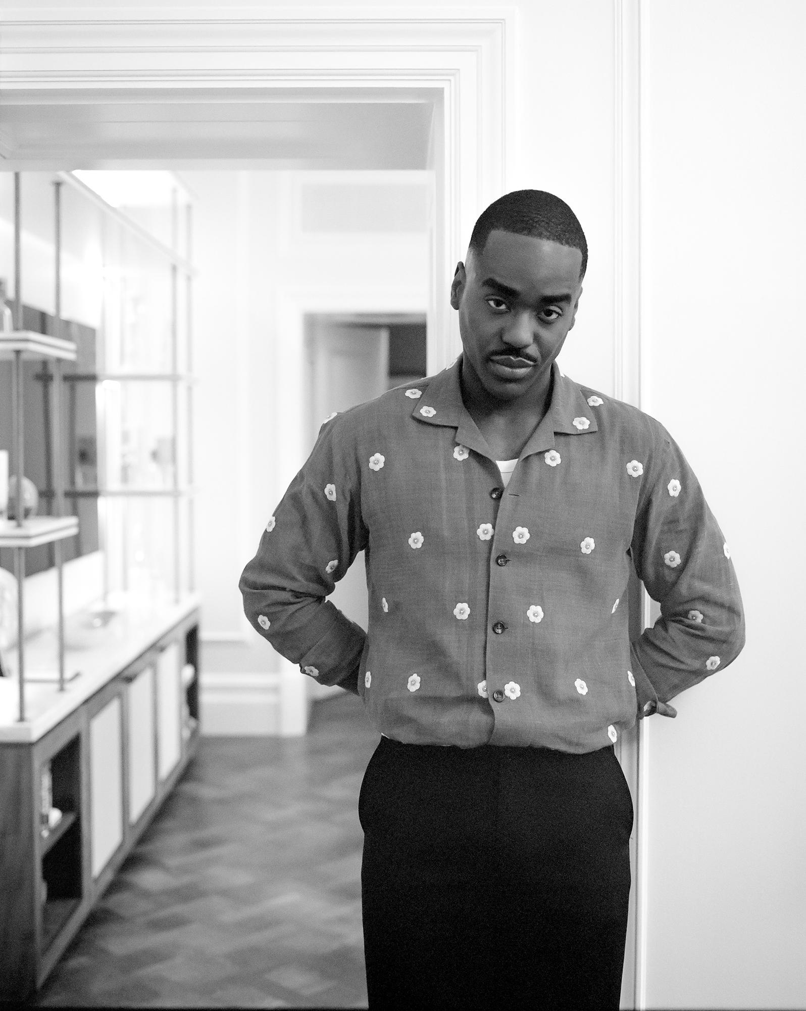 A man in a patterned shirt and trousers standing in a hallway with a white background.