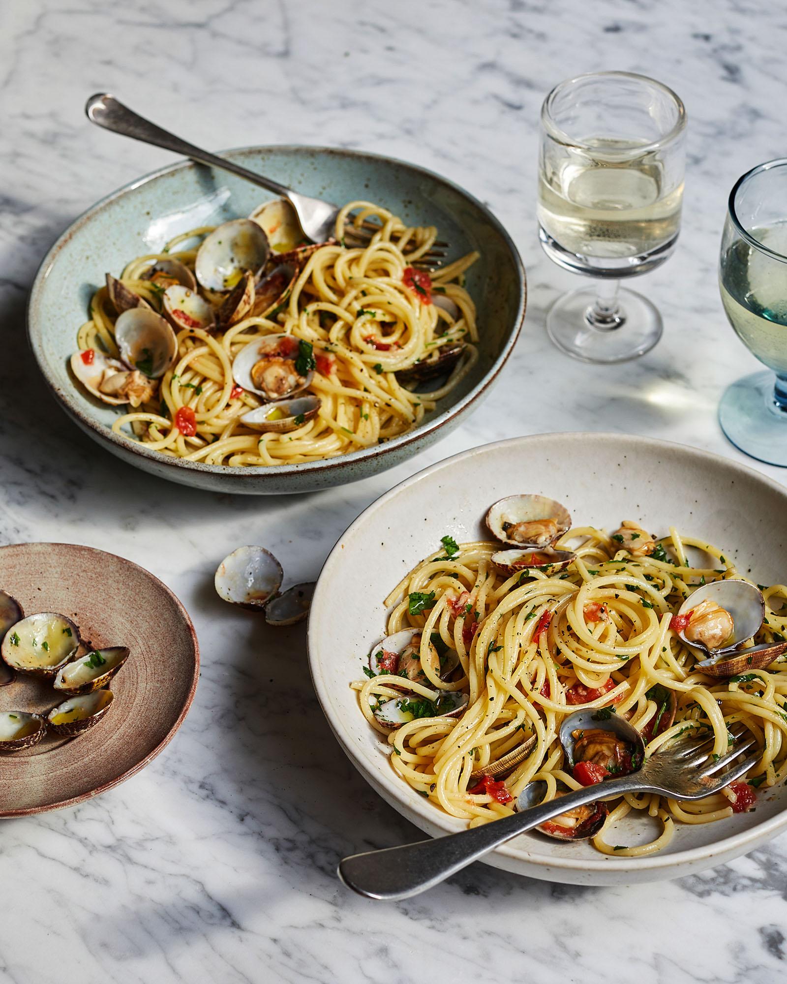 A bowl of spaghetti with clams, served with a fork and a glass of wine on a marble table.