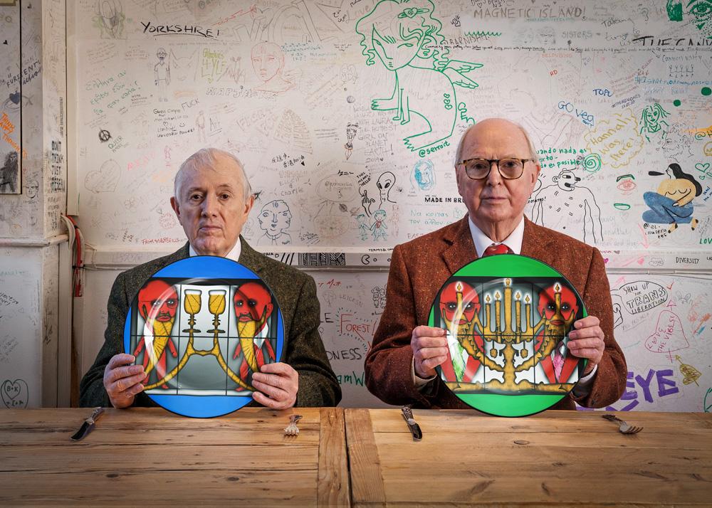 Two older men in suits and ties sitting at a table, each holding a plate with a unique, abstract, and colorful design.