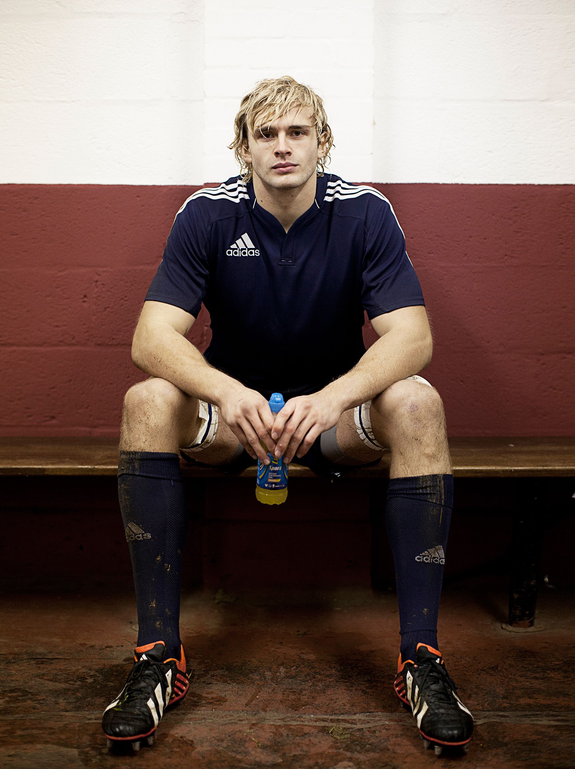 A man in a blue Adidas rugby jersey, sitting on a bench, holding a sports drink bottle.