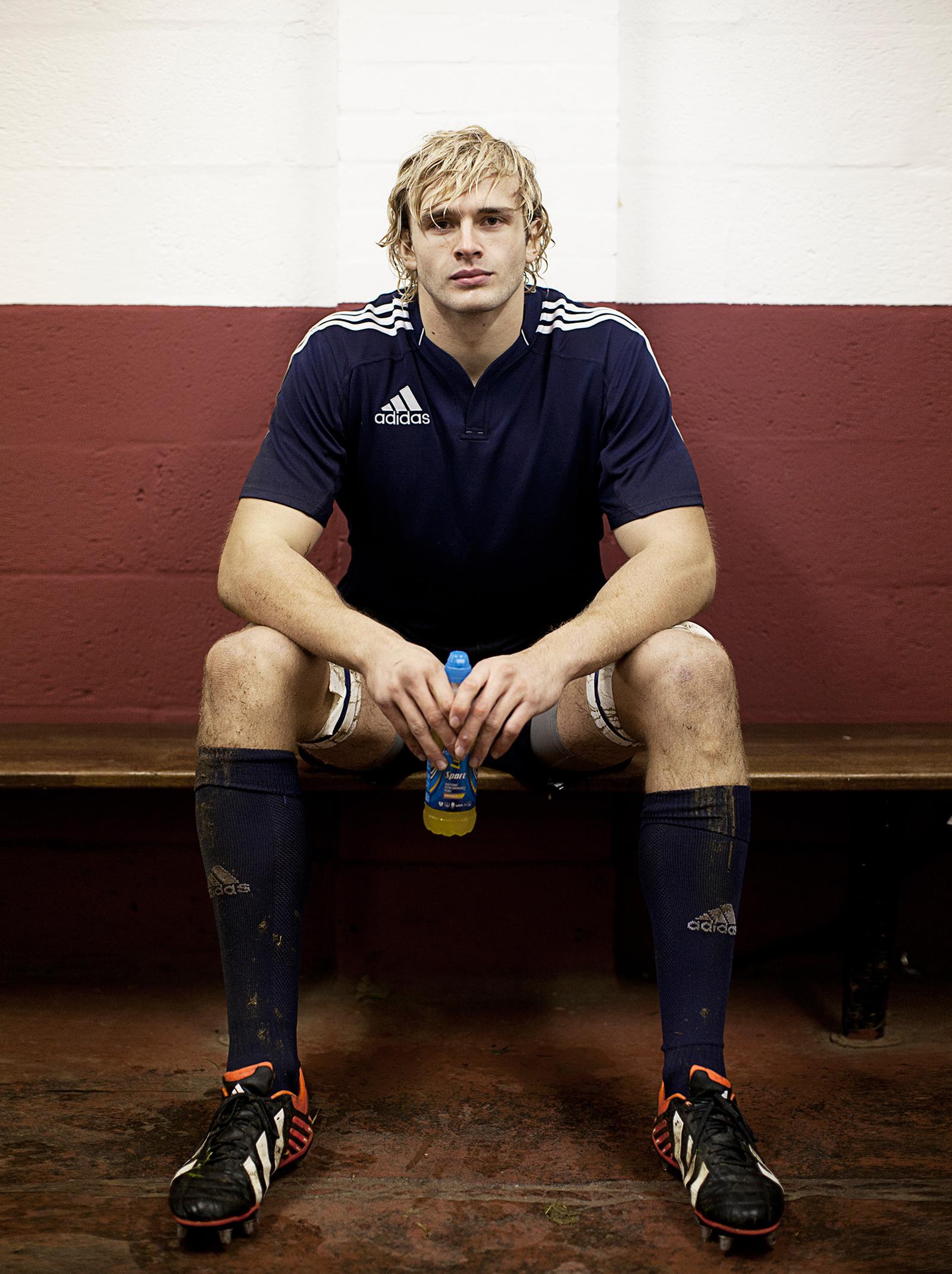 A man in a blue Adidas rugby jersey, sitting on a bench, holding a sports drink bottle.