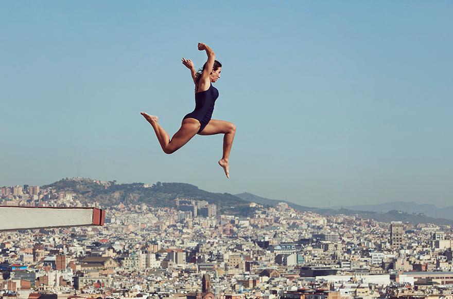 A woman in a black swimsuit is in mid-air, performing a diving or jumping motion over a city skyline.
