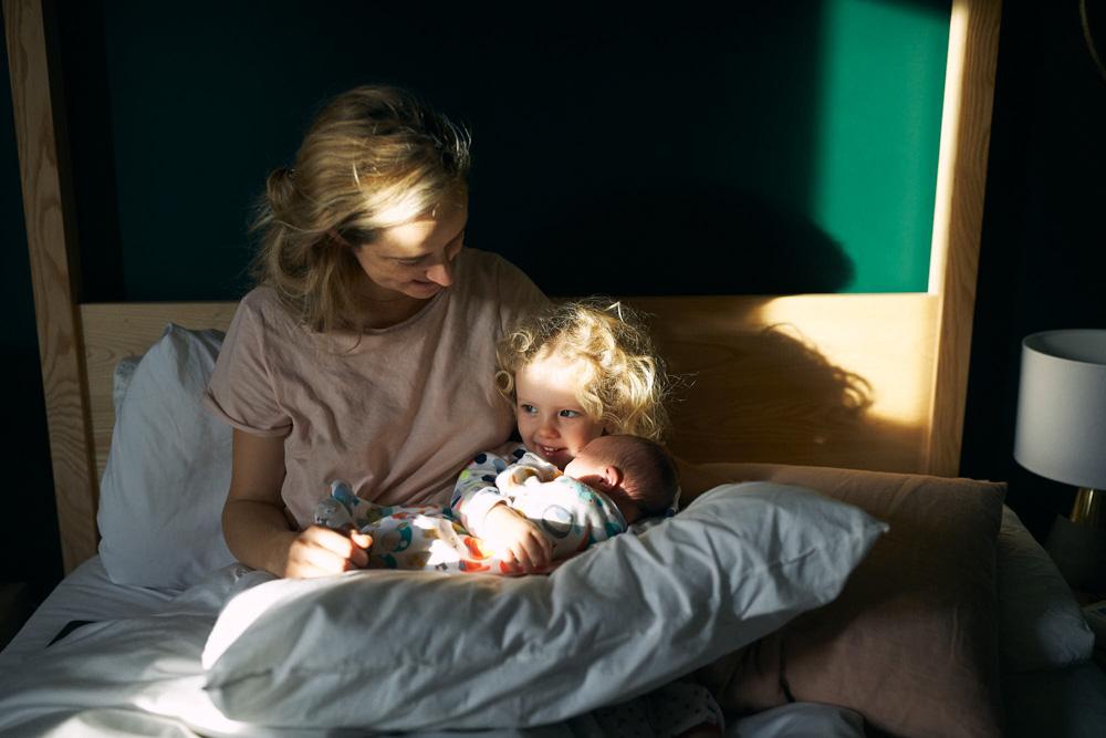 A mother and her two children sit on a bed, surrounded by sunlight. The mother cradles a baby in her arms, while a toddler sits on her lap, both with blonde hair. The atmosphere.