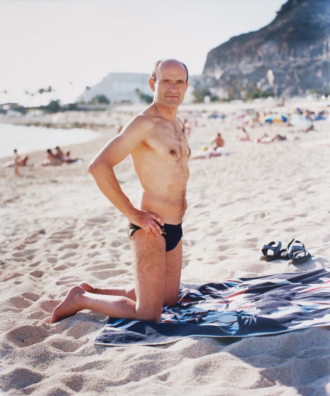 A man in his 40s, wearing a pair of black swim shorts, standing on a beach with his hands on his hips, looking out at the ocean.