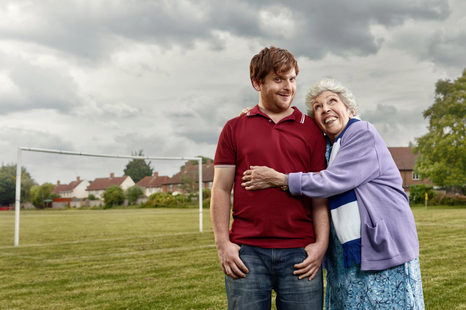 A man in a red polo shirt and jeans standing on a football field with an elderly woman in a blue dress and a purple cardigan, with a goal in the background.