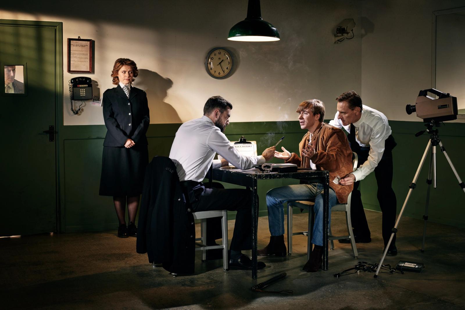 A man in a brown leather jacket is being questioned by two men in white shirts at an office or police station, with a woman, camera, and clock visible.