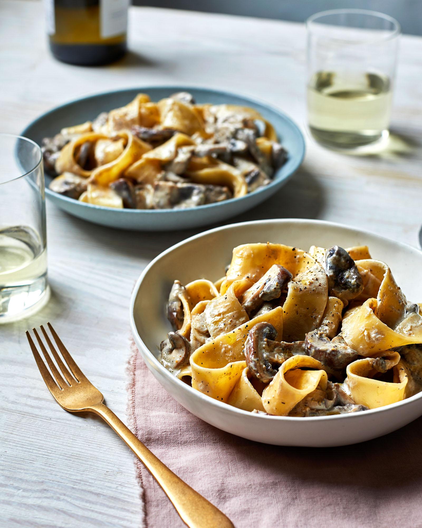 A bowl of creamy pasta with mushrooms, served on a table with a wine glass and a bottle of wine in the background.