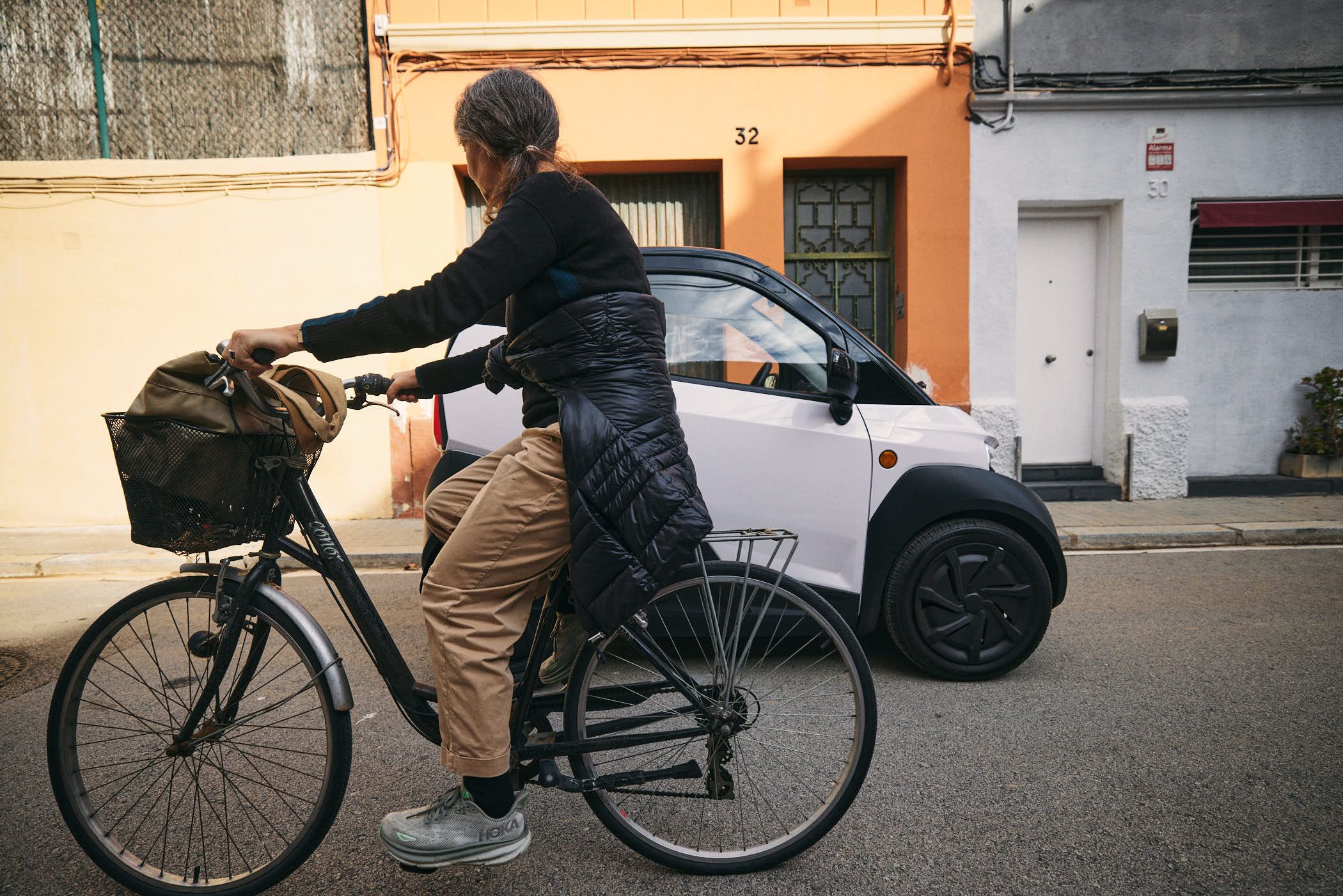 A woman with grey hair rides a bicycle down a street, wearing a black sweater, tan pants, and grey sneakers. She has a brown backpack on the front of her bike and a black jacket.