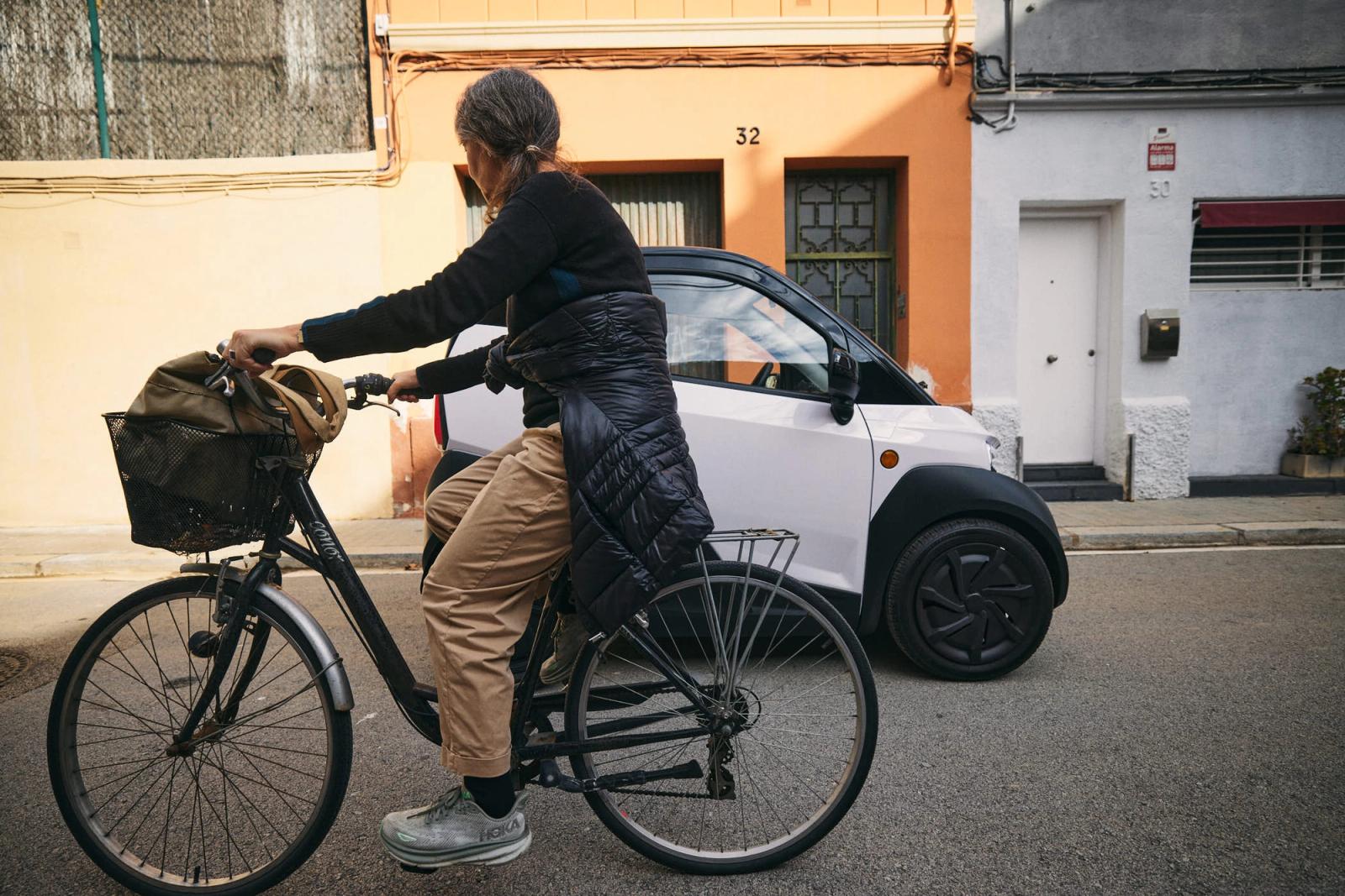 A woman with grey hair rides a bicycle down a street, wearing a black sweater, tan pants, and grey sneakers. She has a brown backpack on the front of her bike and a black jacket.