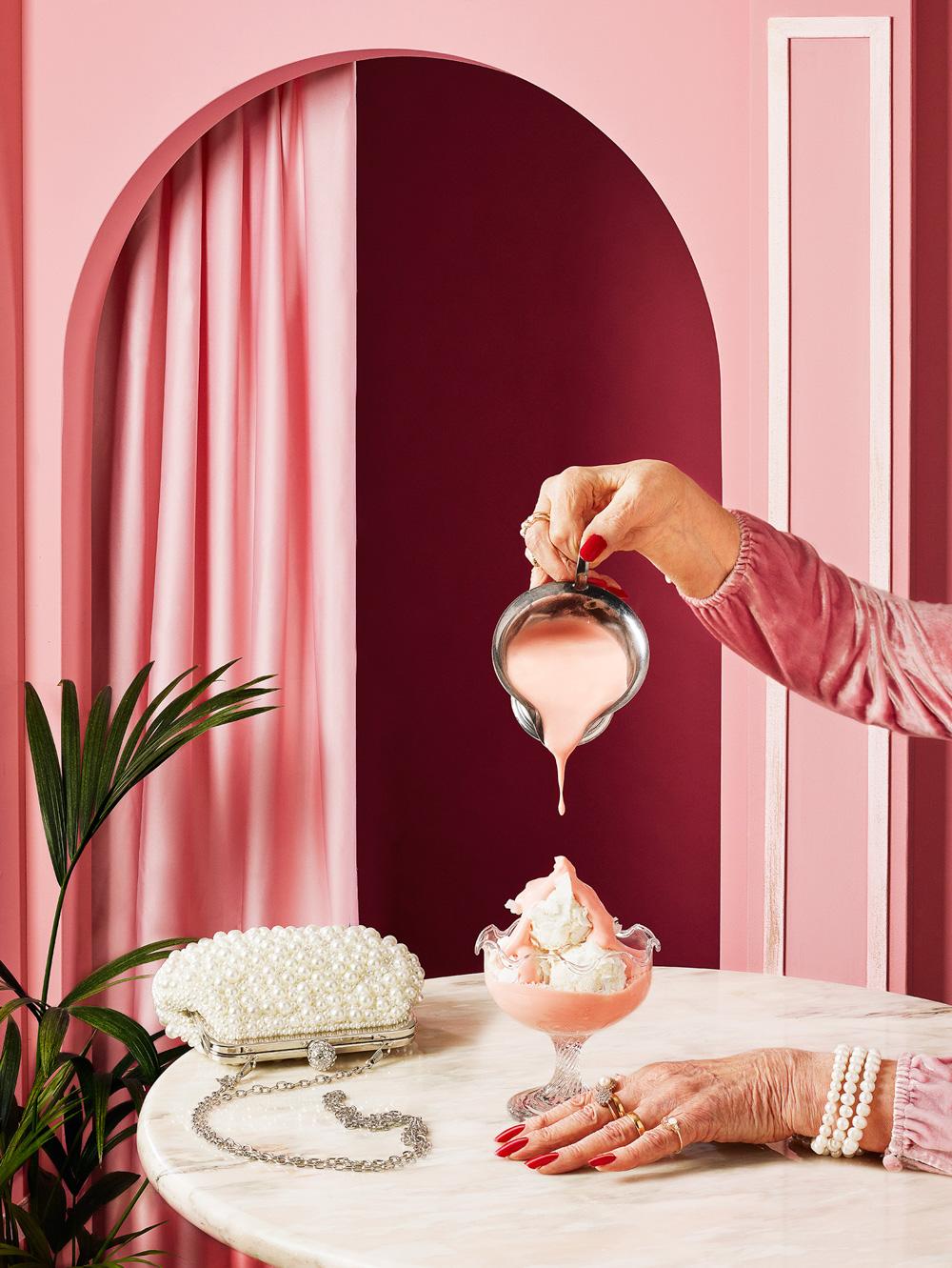 A woman's hand pouring a pink liquid into a glass of ice cream, with a white purse and a plant in the background.