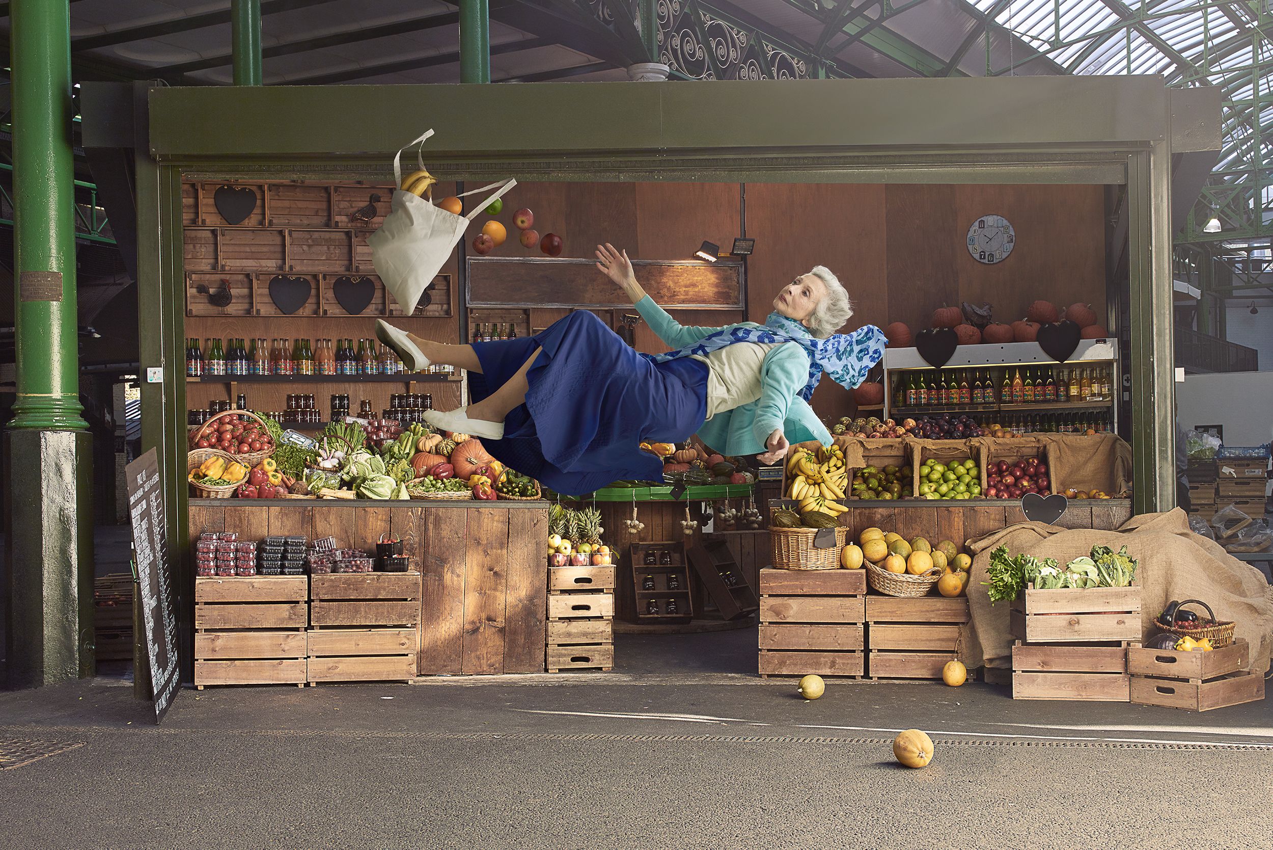 A woman in a blue dress and white shoes is lying on top of a fruit and vegetable stand, surrounded by various types of fruits and vegetables.