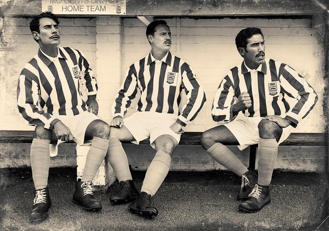 Three men in football jerseys and shorts sitting on a bench, looking at something in the distance.