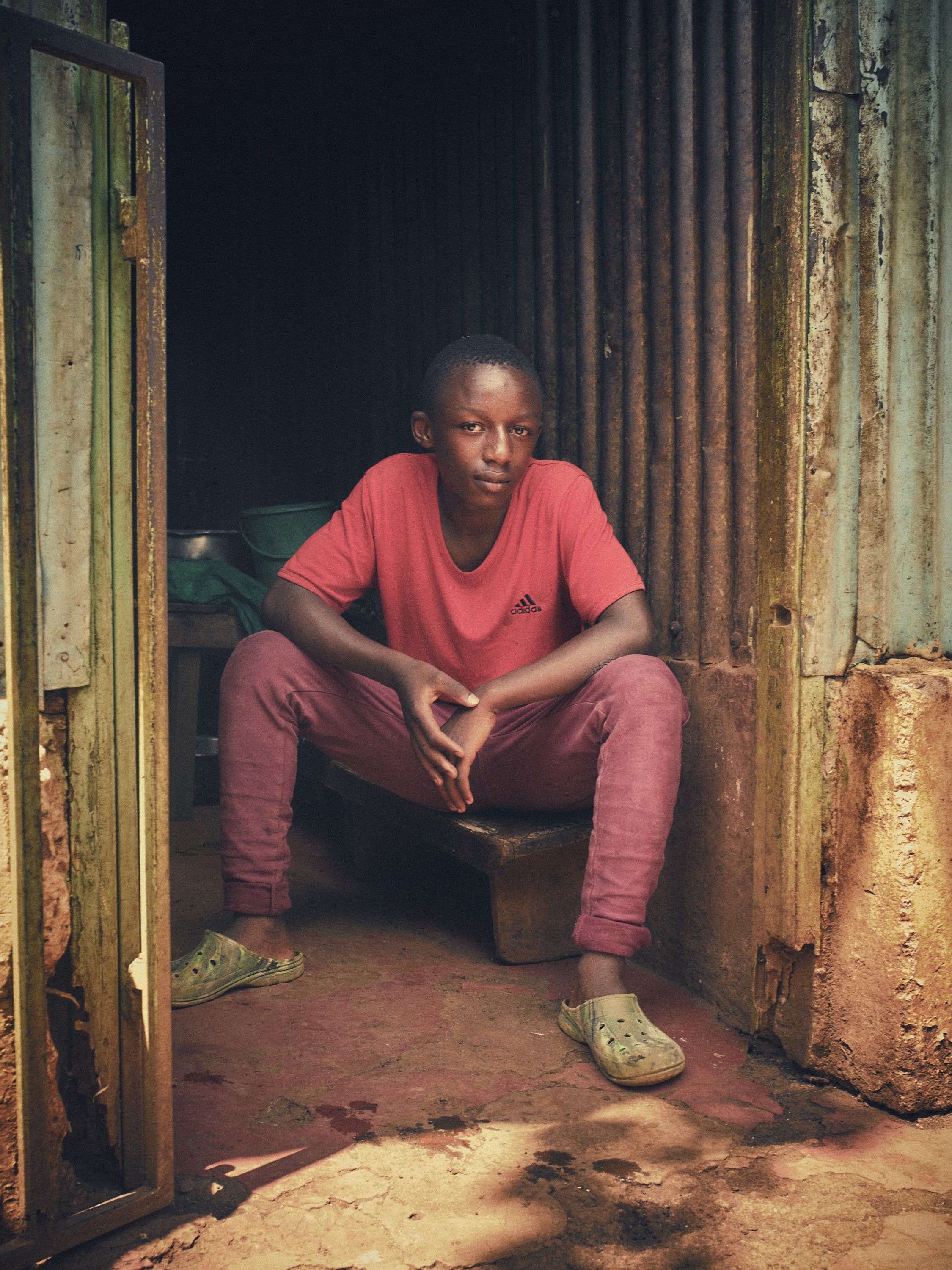 A young boy in a red t-shirt and pink pants sitting on a wooden stool in a dimly lit, rustic outdoor setting.