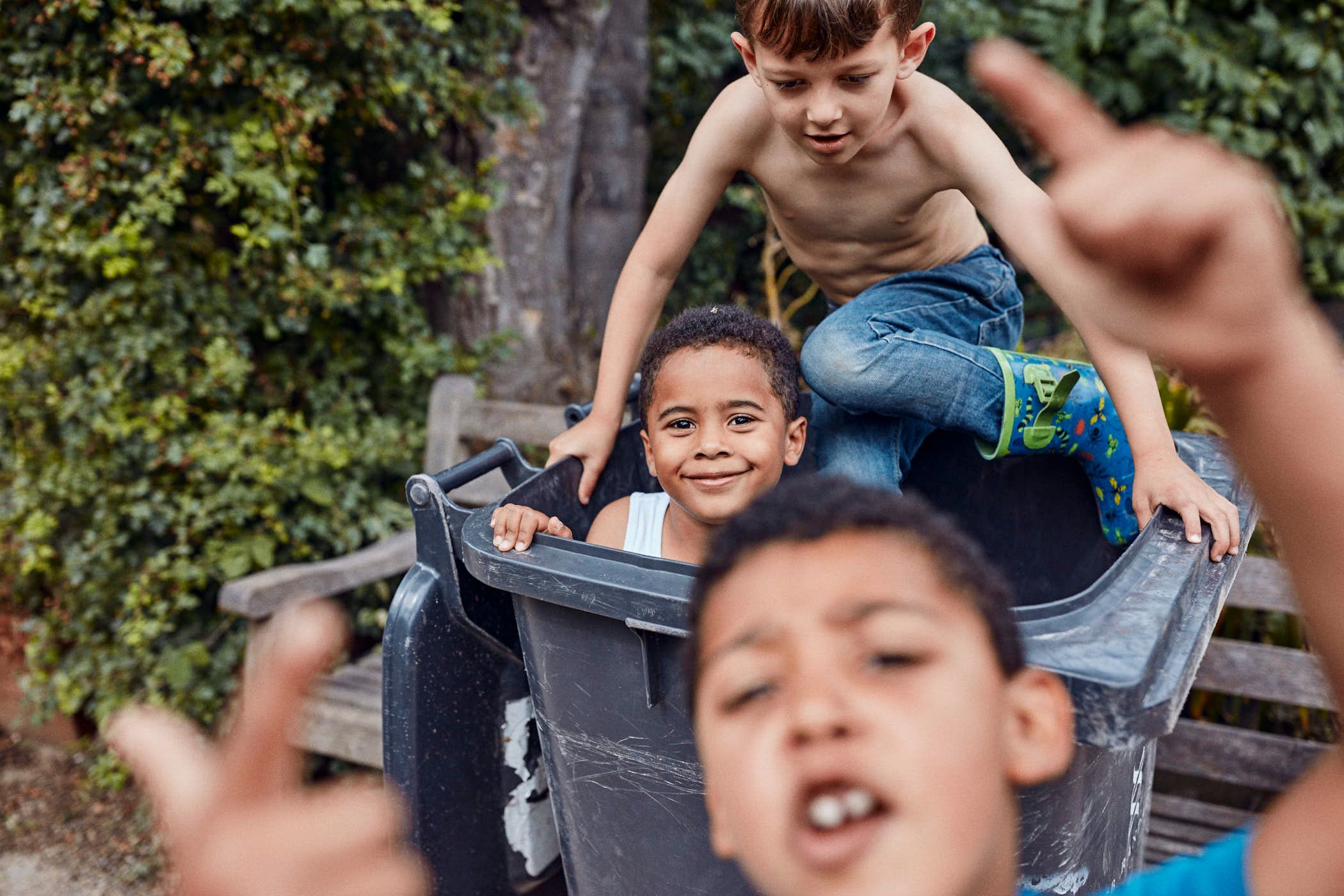 A group of children playing in a large trash bin, with one boy perched on top of the others. The atmosphere is playful and joyful, with the children's faces and body language.