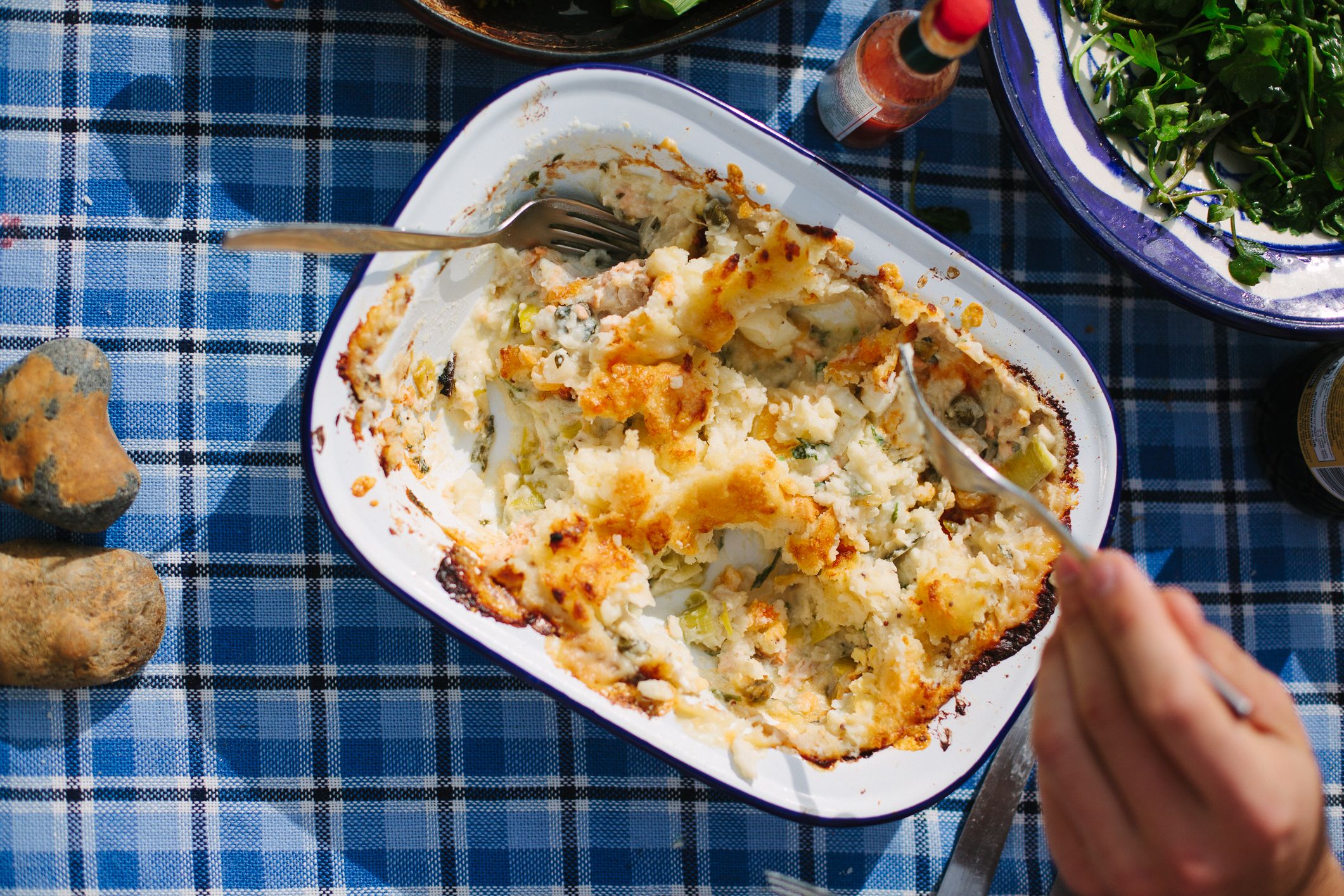 A person is eating a dish of mac and cheese from a white, blue-rimmed, rectangular baking dish, with a fork in their right hand, on a blue and white checkered tablecloth.